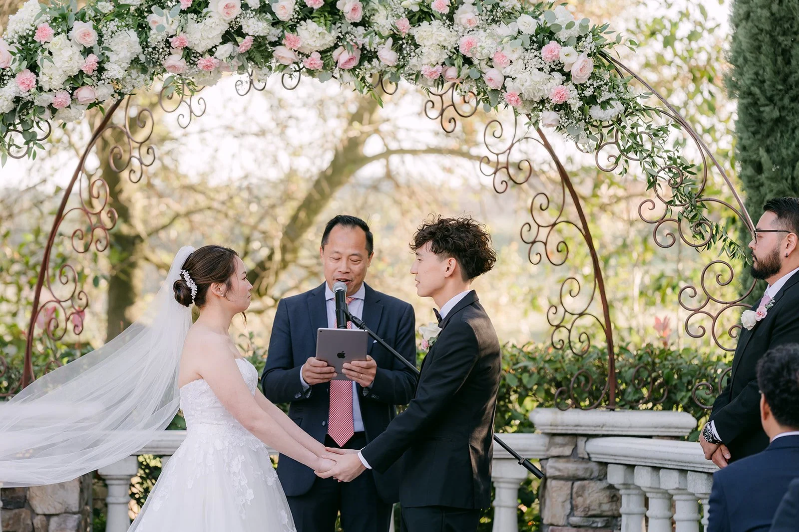 Victoria and Tony holding hands under the floral arch as the pastor leads the ceremony