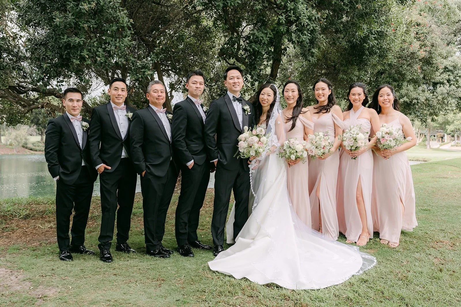 The full wedding party in black tuxedos and dusty pink gowns on the lawn