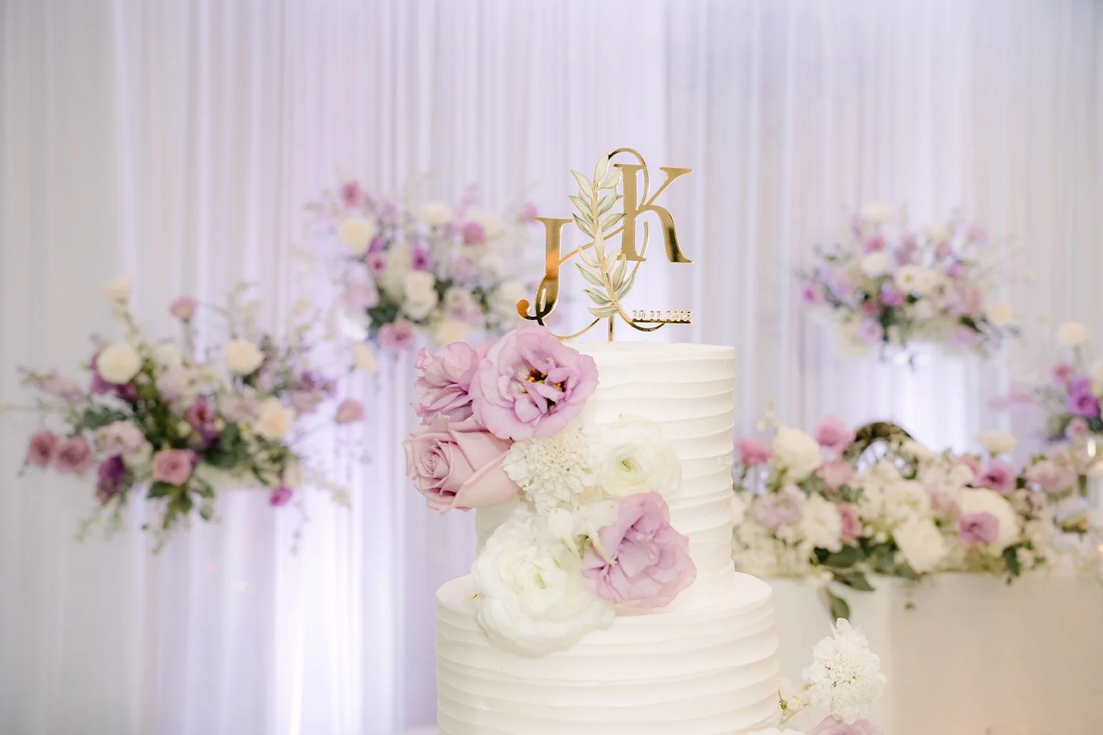 The white tiered cake topped with a gold JK monogram and lavender roses