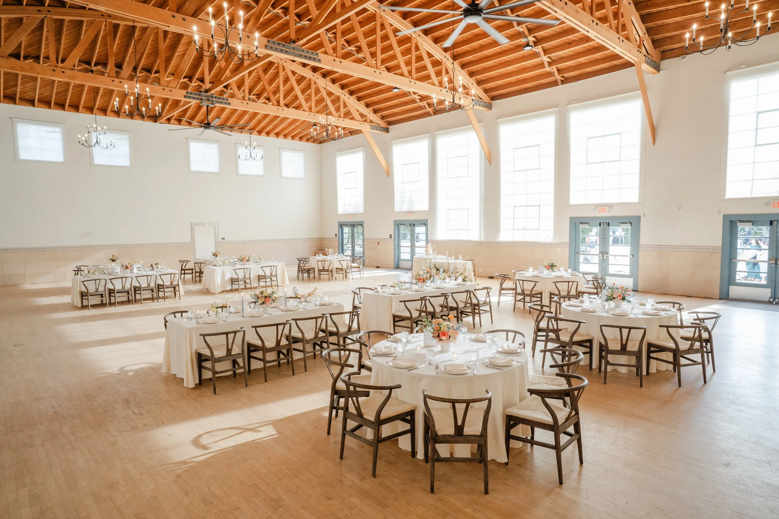 An event hall with high ceilings featuring exposed wooden beams, chandeliers, and large windows. The room is decorated with round tables covered in white tablecloths, floral centerpieces, and place settings, likely prepared for a wedding or celebrati