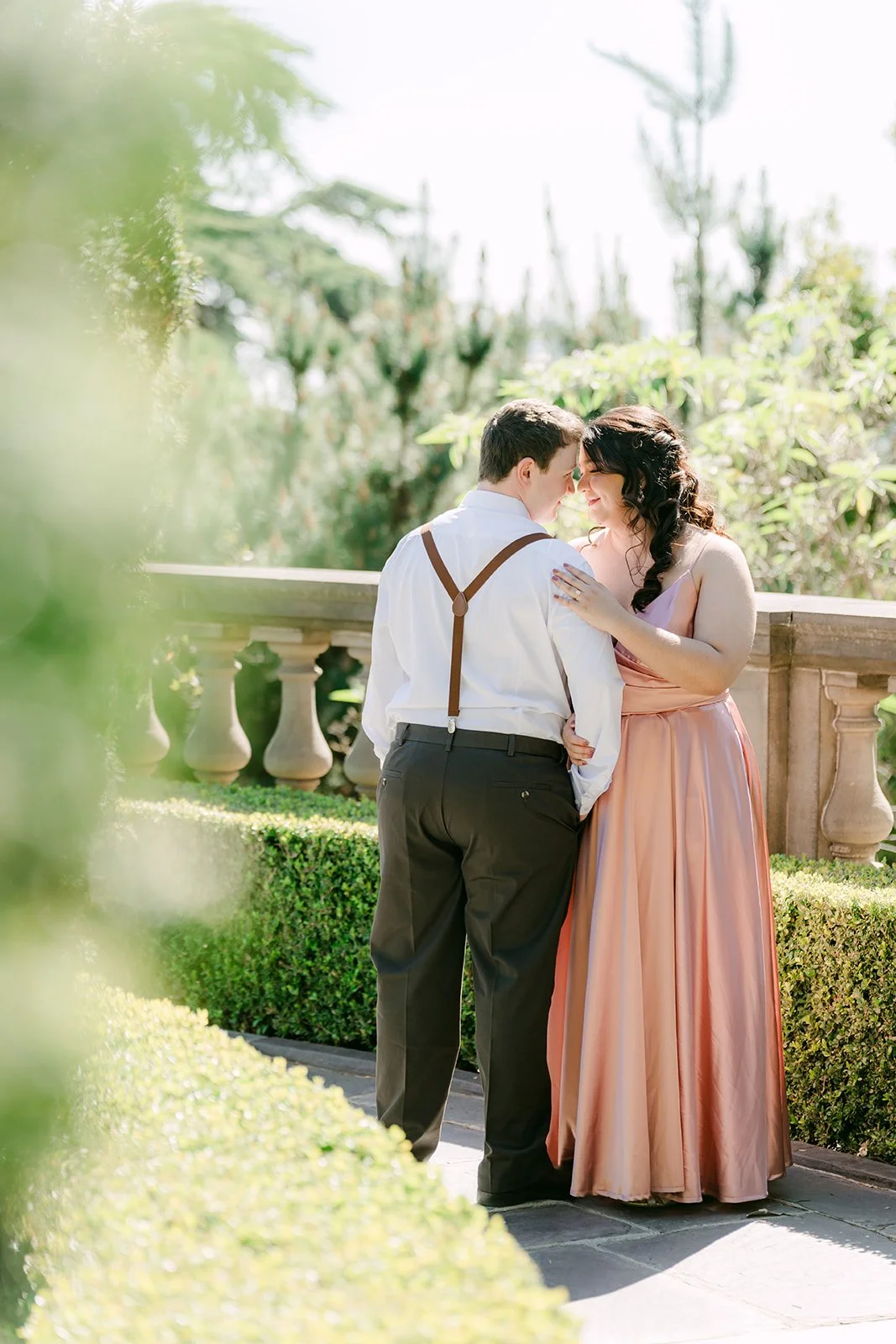 Saira and Matthew forehead to forehead beside the garden hedges.