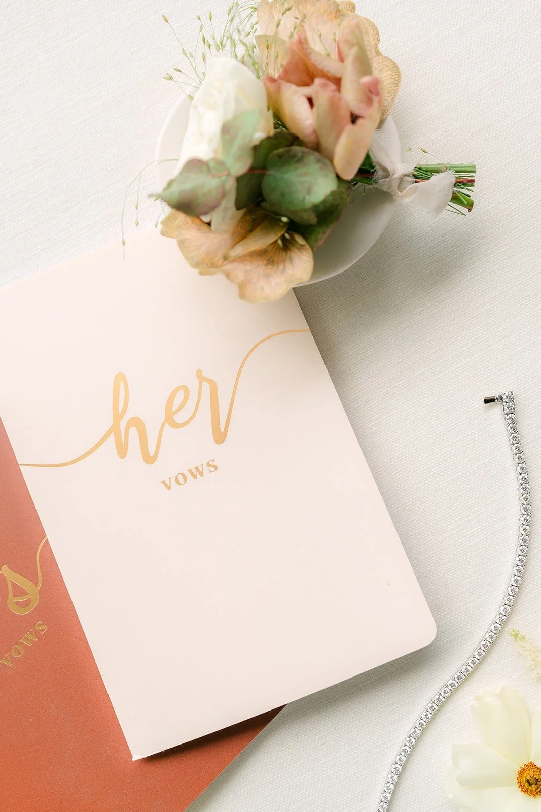 A vow book marked "her" with a blush rose boutonniere and a sparkling bracelet