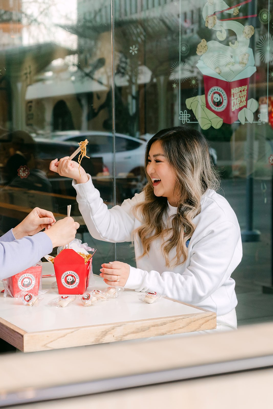 Linh offers Ankush a bite with chopsticks at a window table.