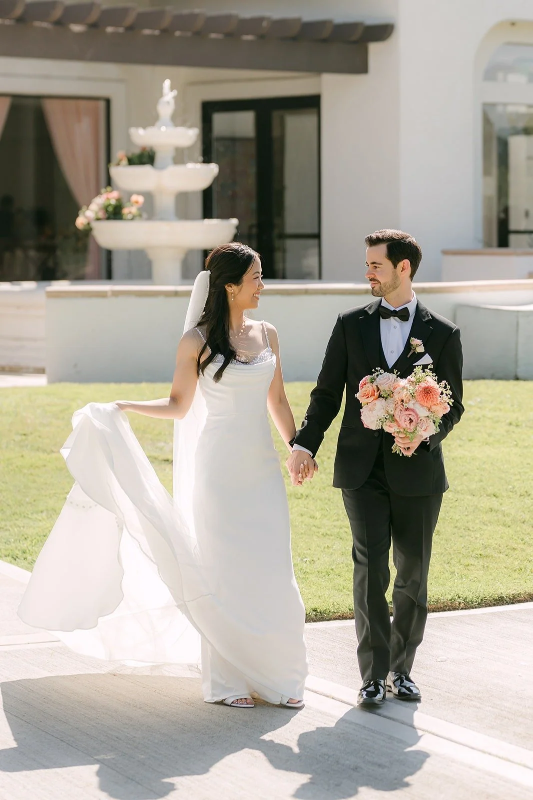 Stephanie and Kevin walking hand in hand across the courtyard as her veil trails behind
