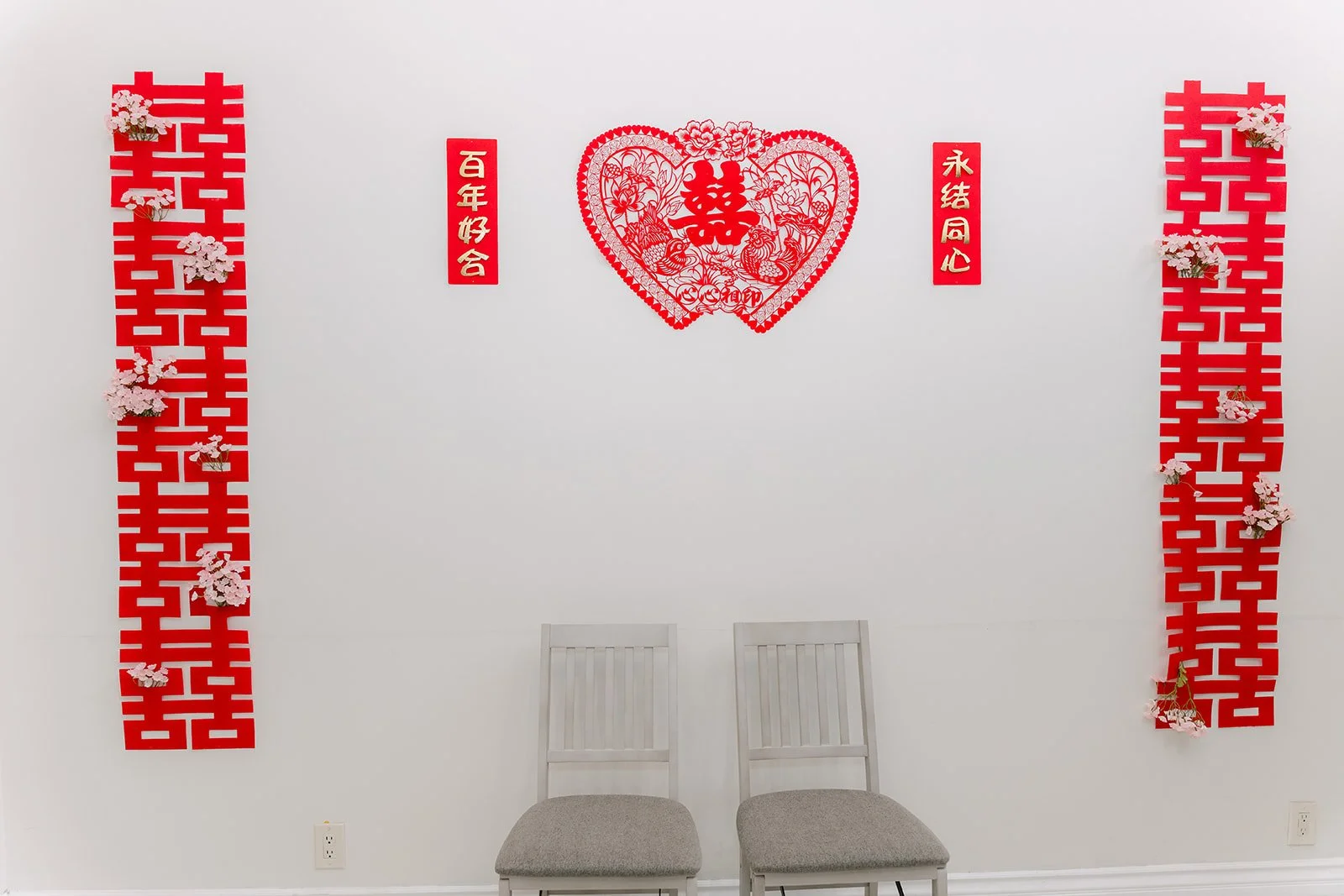 Heart shaped double happiness papercut and red couplets on a white wall above two chairs for the elders.