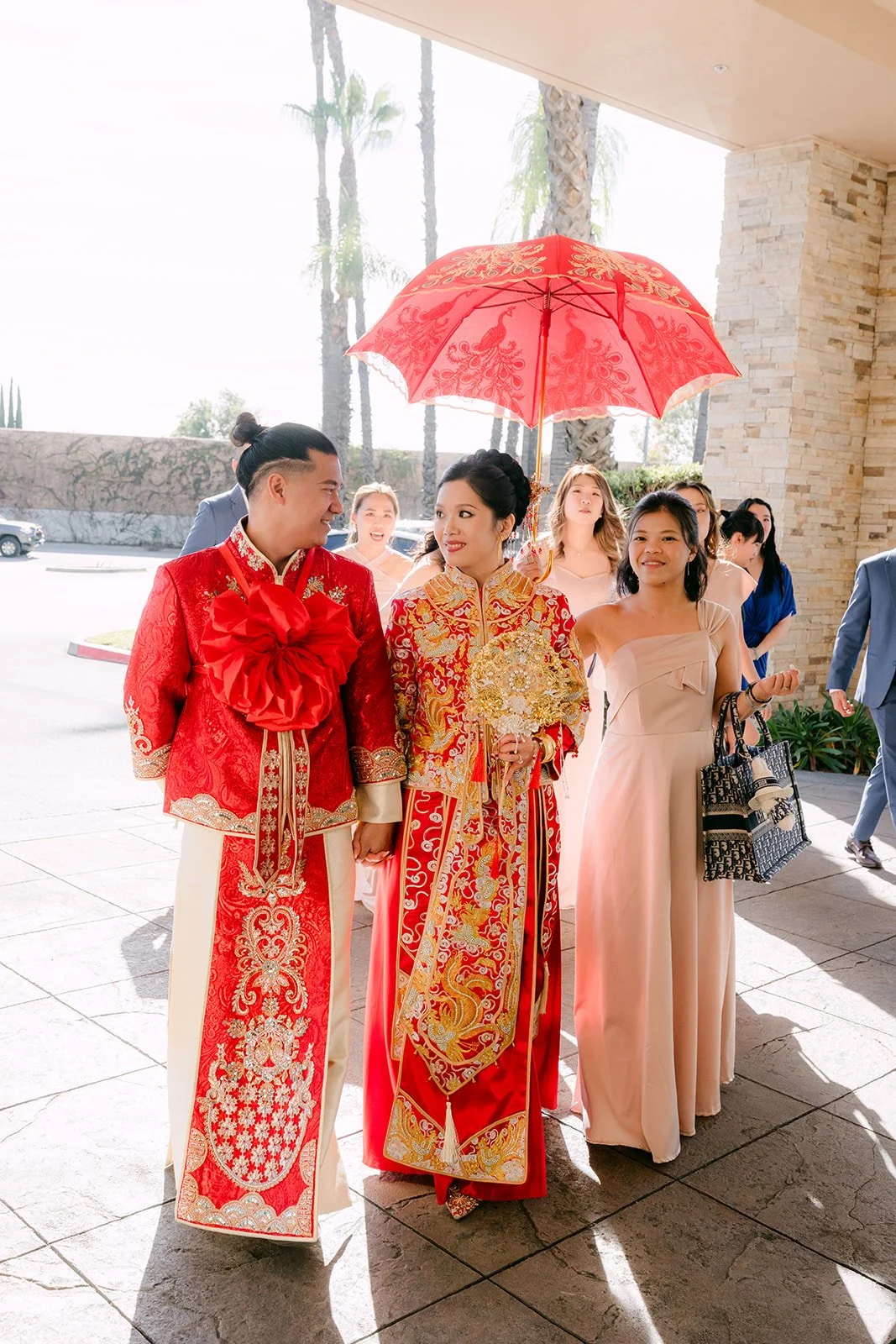 The bride and groom walking out under a red umbrella with family following.