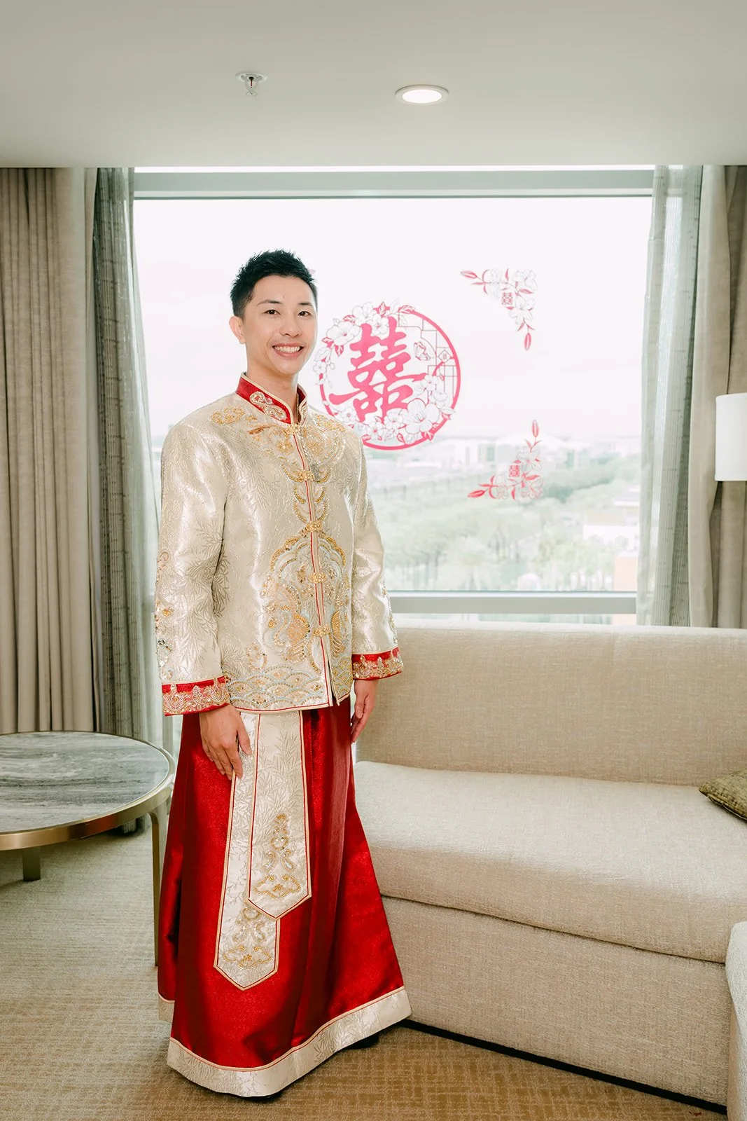 A groom in a cream and red changshan standing by the window.