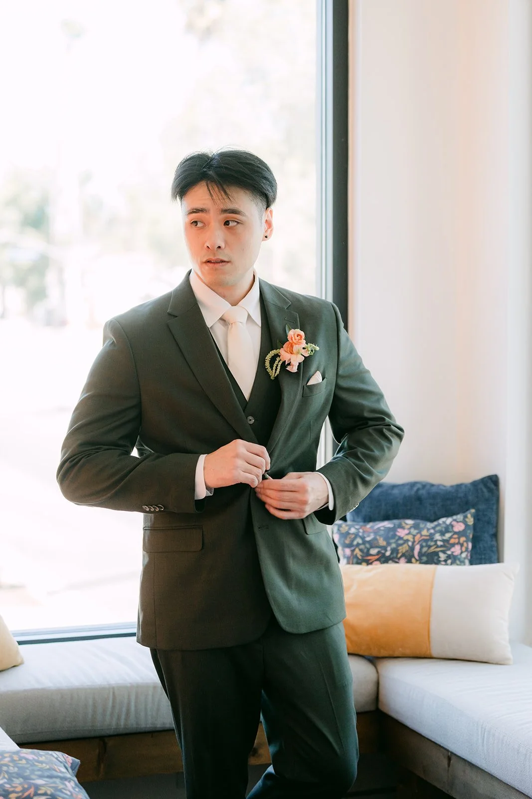 Alex buttoning his olive suit jacket by the window