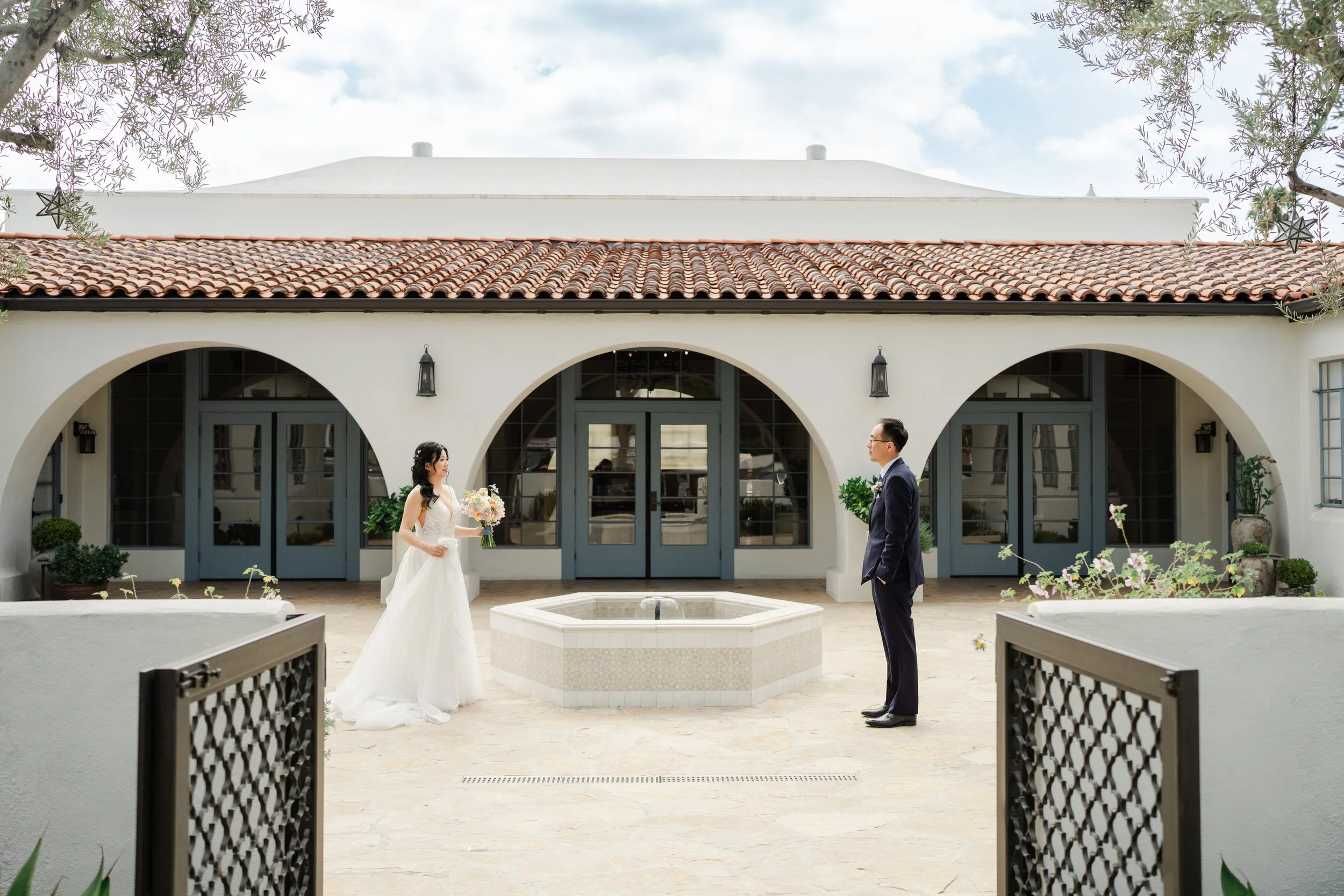A bride and groom stand facing each other in an outdoor courtyard of a white building with Spanish-style arched doors and a tiled roof. The bride wears a white wedding dress and holds a bouquet of flowers, while the groom wears a dark suit and glasse