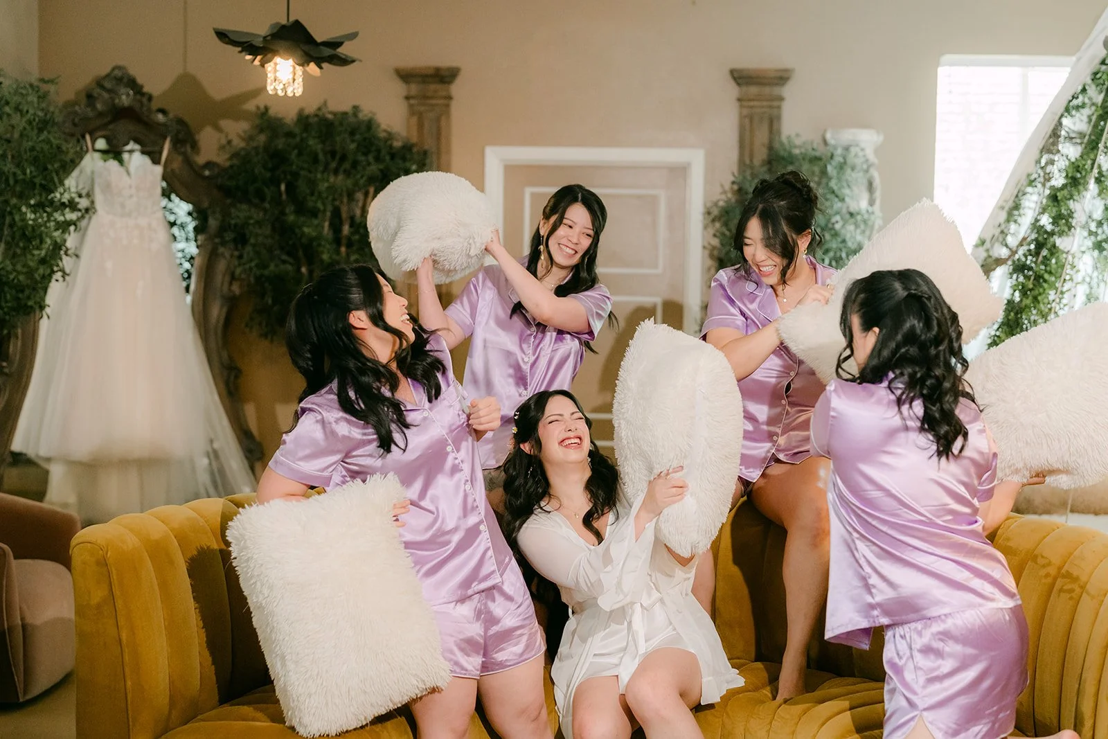 Bridesmaids in a pillow fight with the bride on the mustard sofa