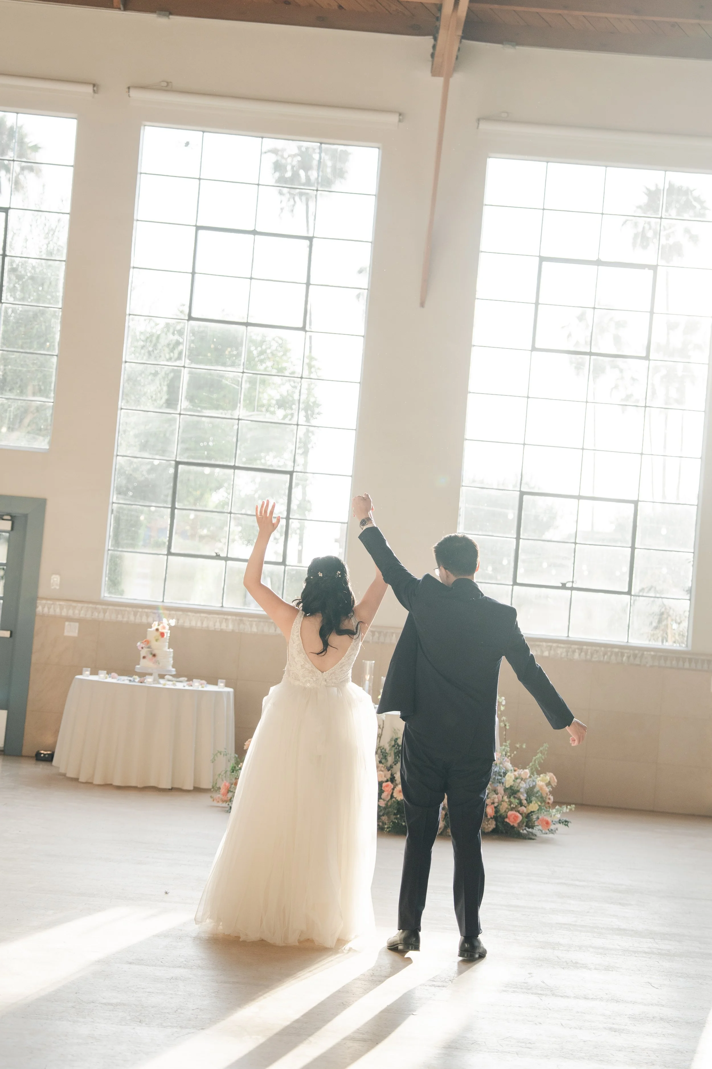 A bride and groom are holding hands and raising them in celebration, standing in a sunlit indoor venue with large windows. The bride wears a white lace dress and the groom wears a black suit. A wedding cake and flowers are visible in the background.