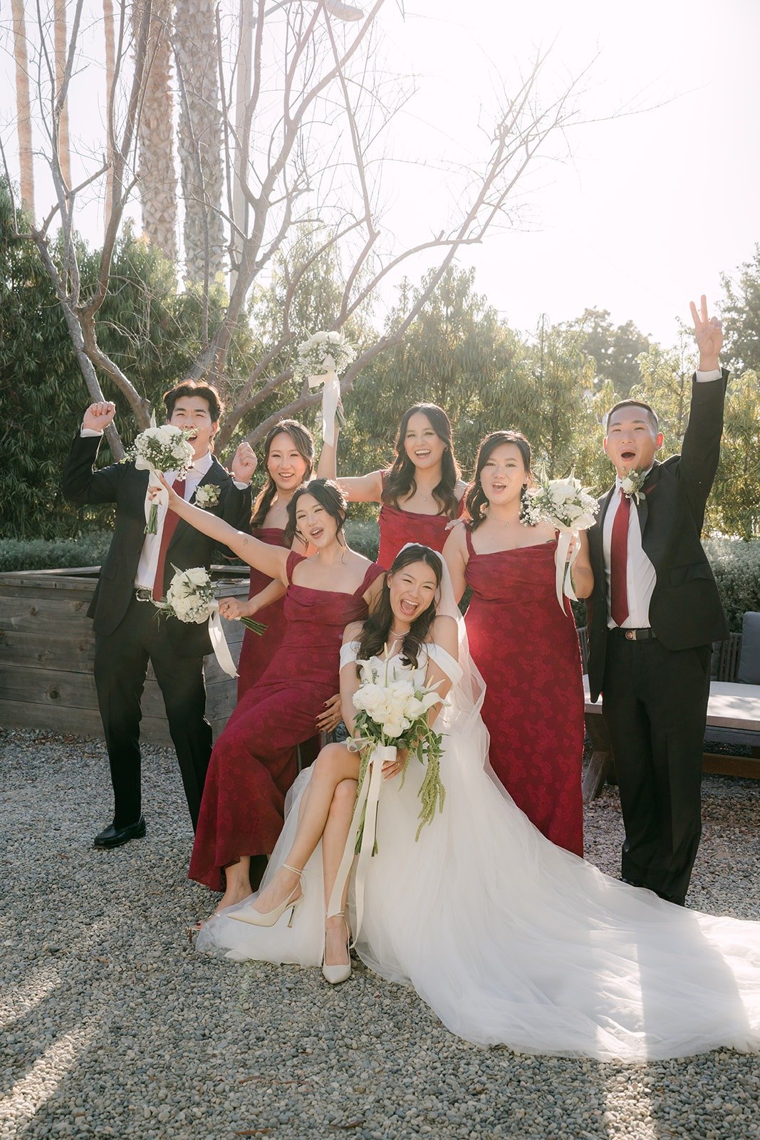 A playful wedding party photo with Jadee seated on the gravel, photographed by One and Only Production