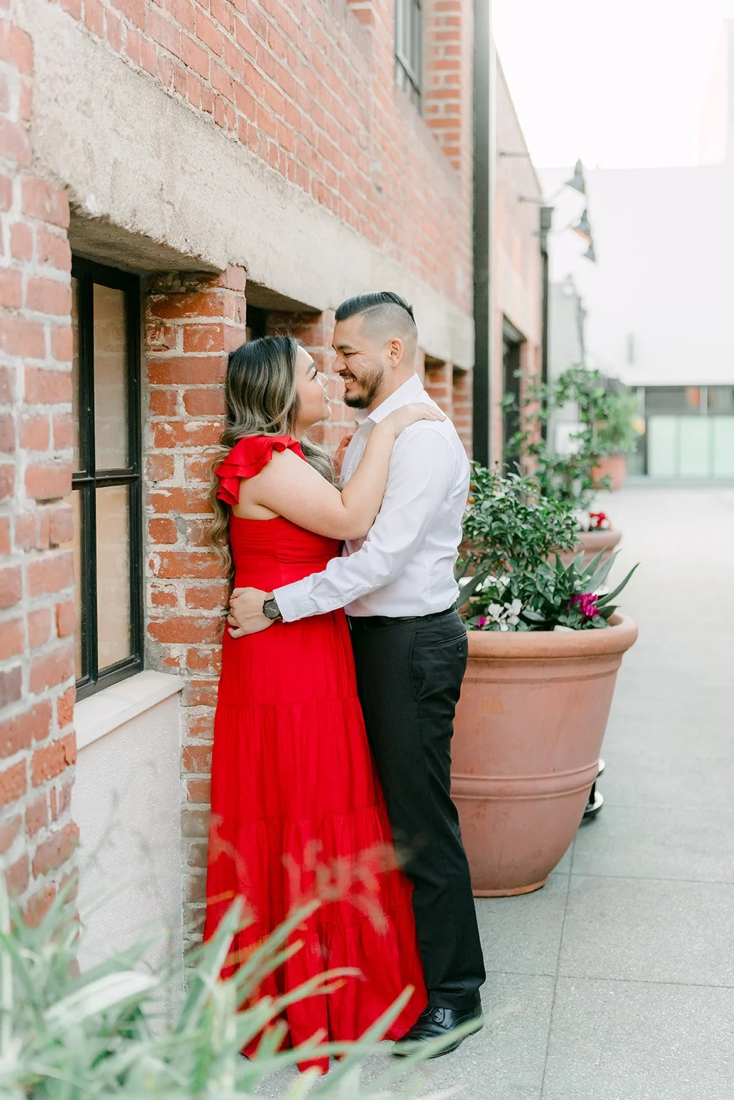 Linh and Ankush embracing beside a red brick wall and potted plants.