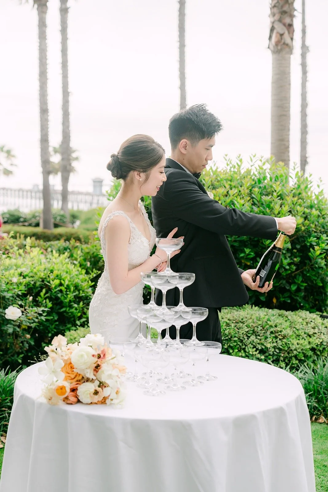 Eli opening a bottle of champagne beside Angela at the champagne tower