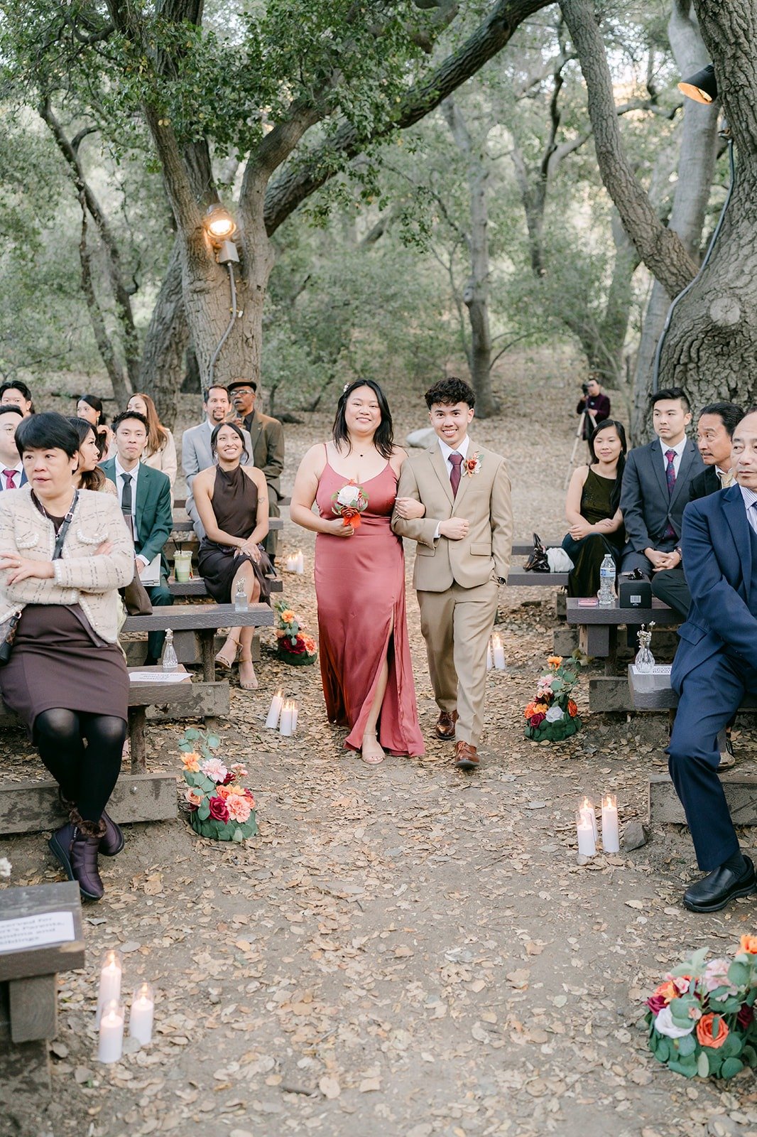 A bridesmaid and groomsman walk down the aisle between the oak trees