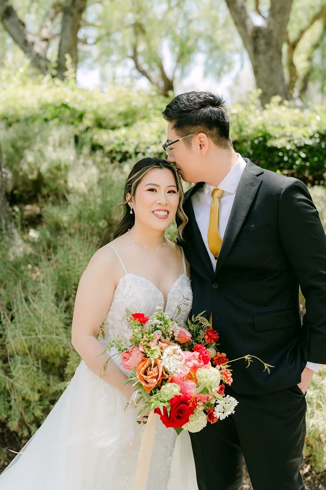 Joanne leaning into Zach with her red and pink bouquet among the trees at Rancho Bernardo Inn
