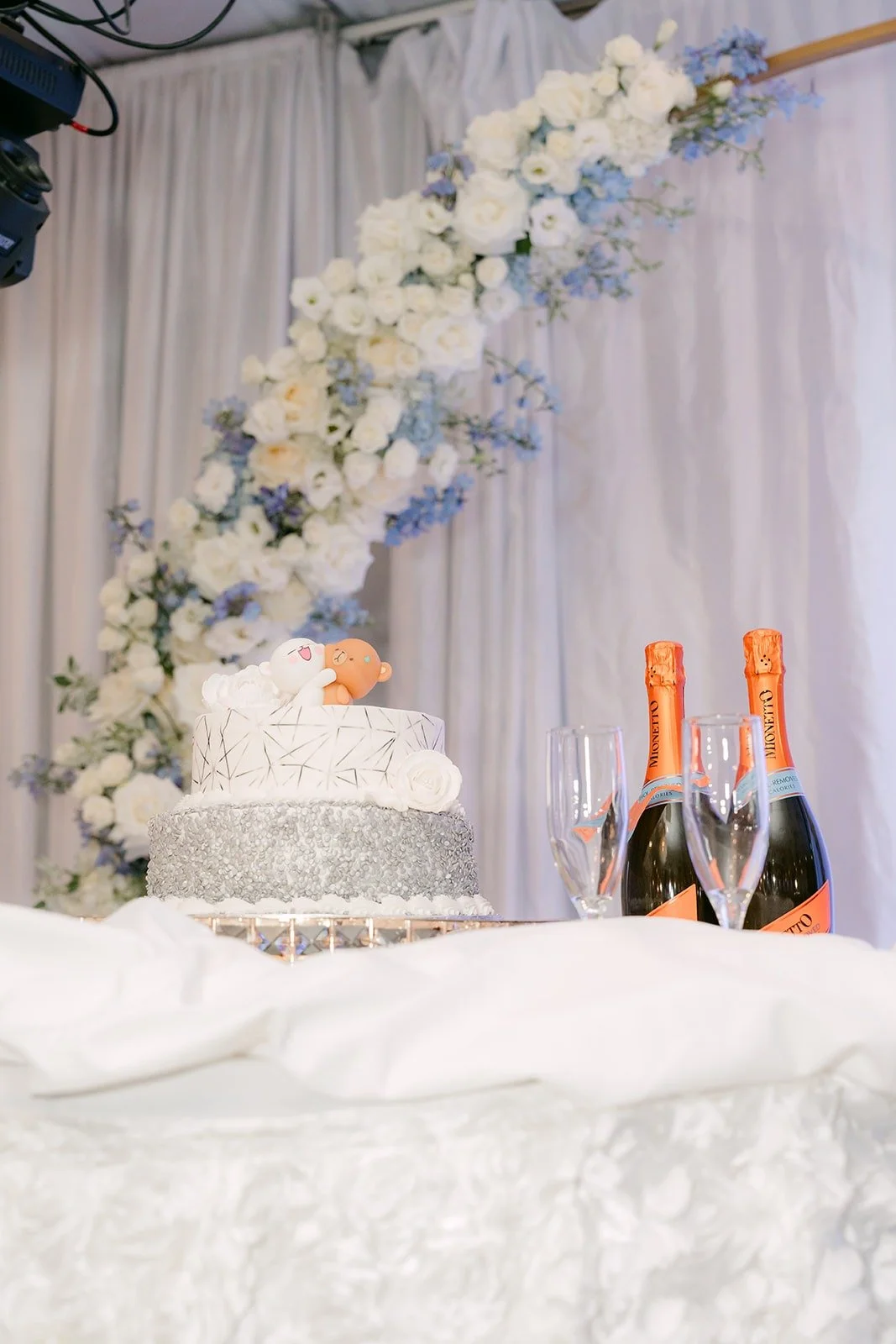 White cake with a sparkling silver tier beside the floral arch