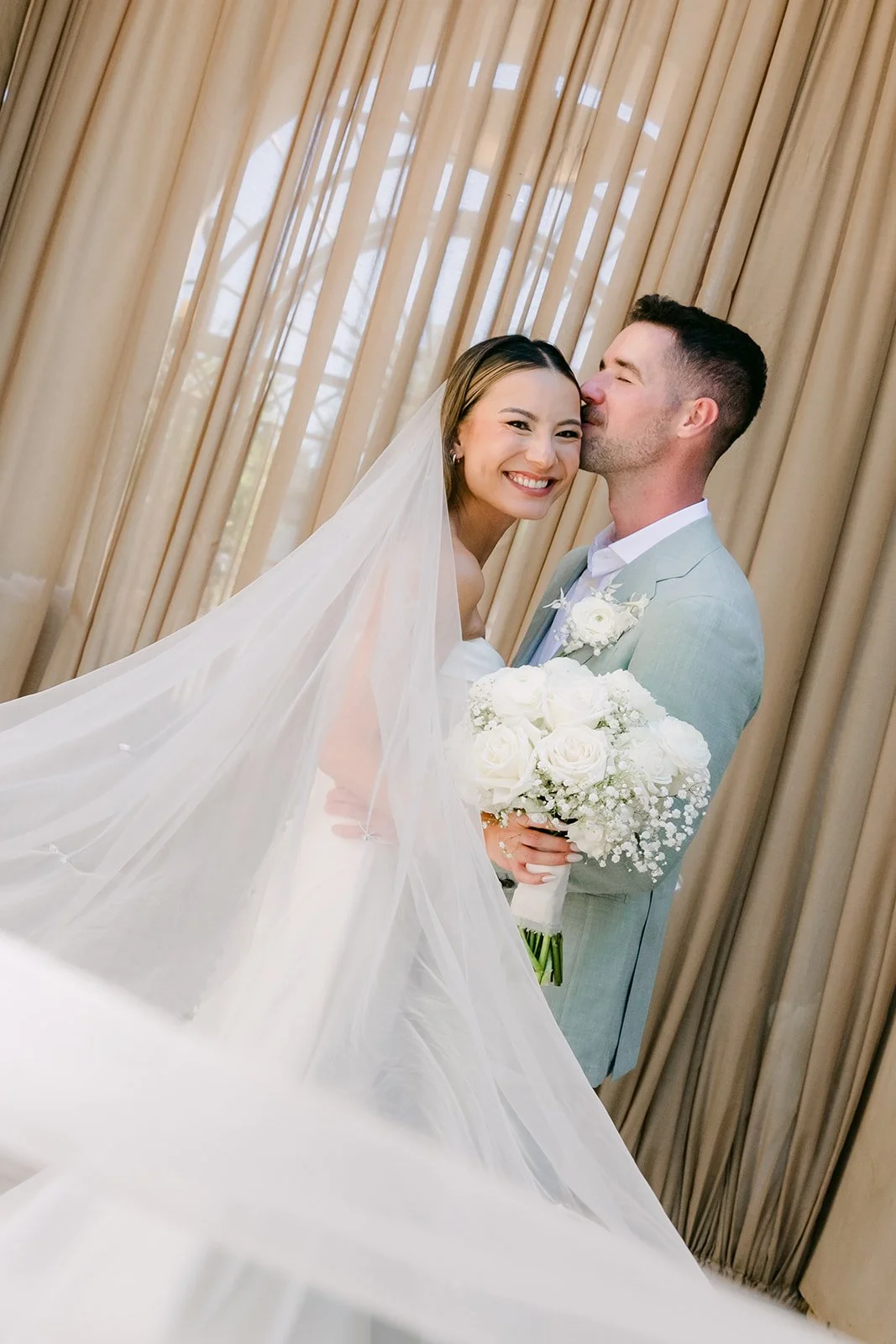 Groom kissing the bride's head as her veil floats in the breeze