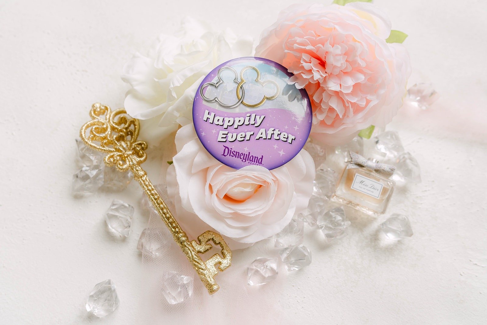 A purple Happily Ever After button and a gold key among pearls and blush roses
