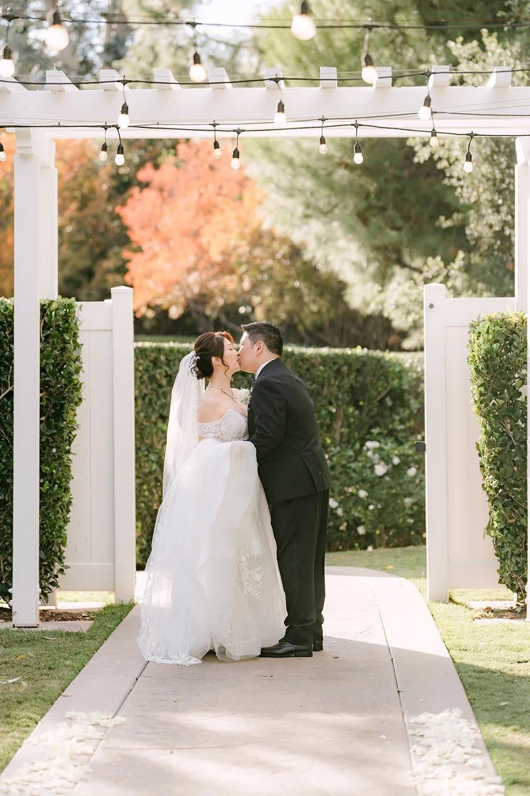Sochi and Jay kiss under the arbor with autumn trees behind them