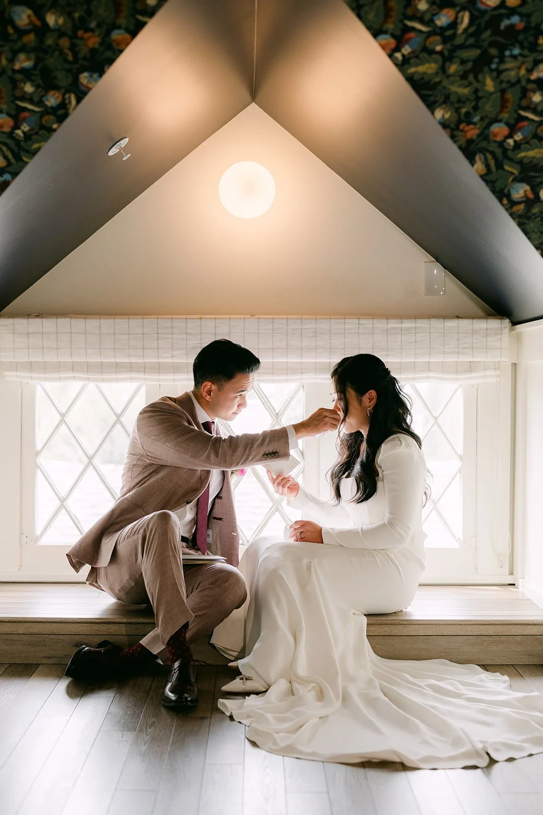 Michael wiping a tear from Denise's cheek in a window nook under a peaked ceiling.