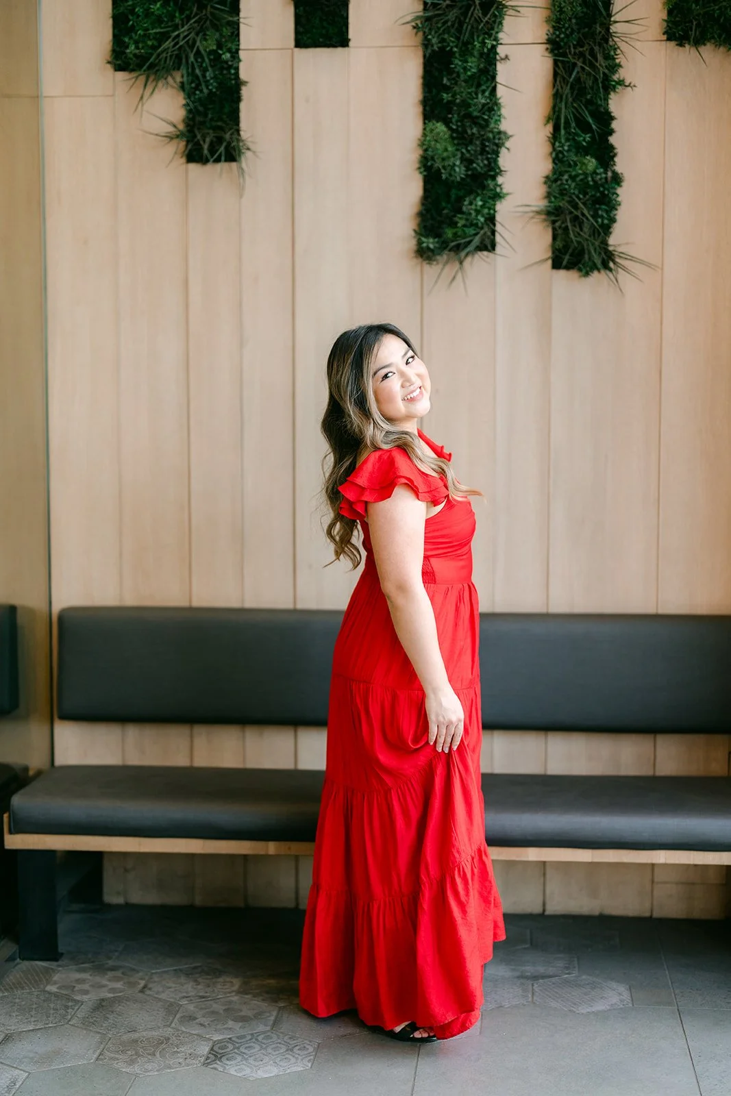 Linh in her red dress in front of the wood wall and hanging plants.