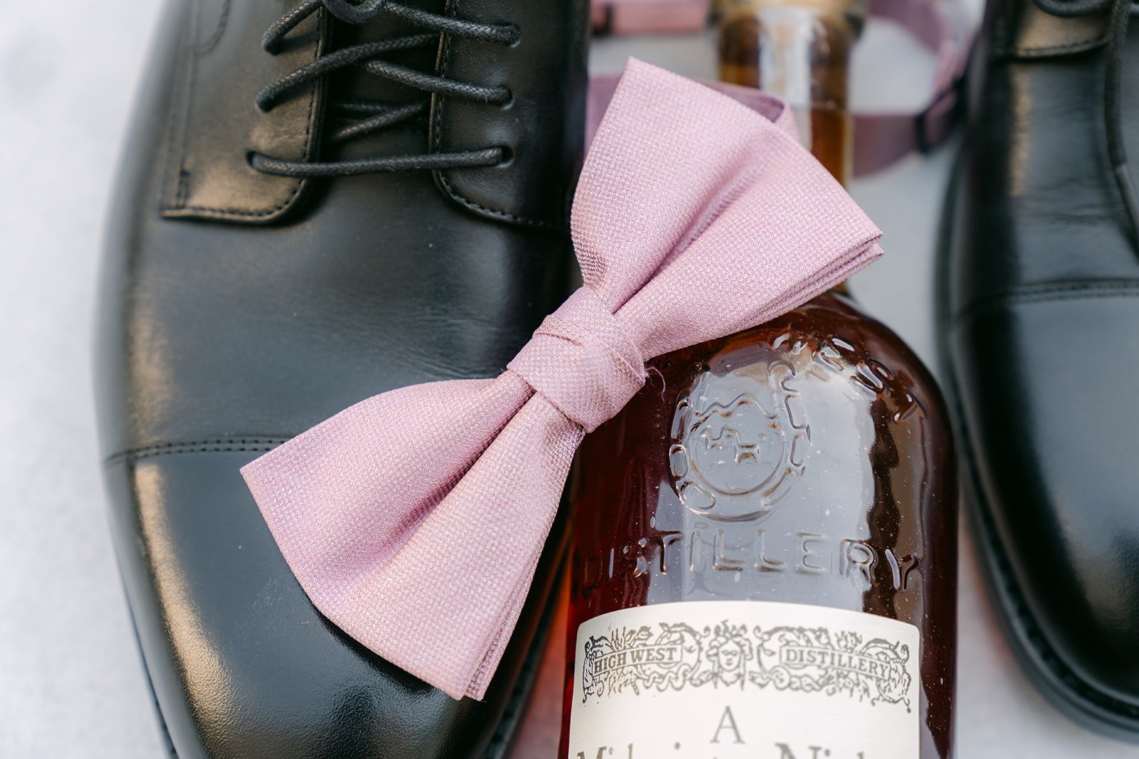 A pink bow tie resting on a bottle of whisky between Brandon's black shoes