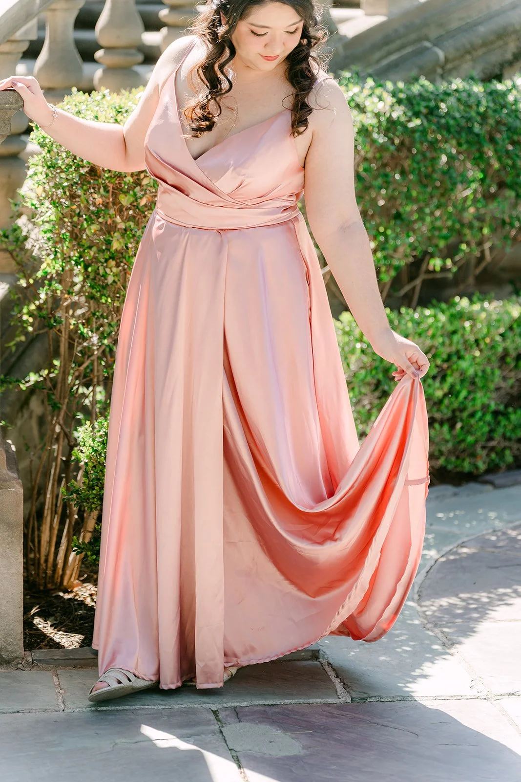 Saira holding out the skirt of her satin gown against the garden hedges.