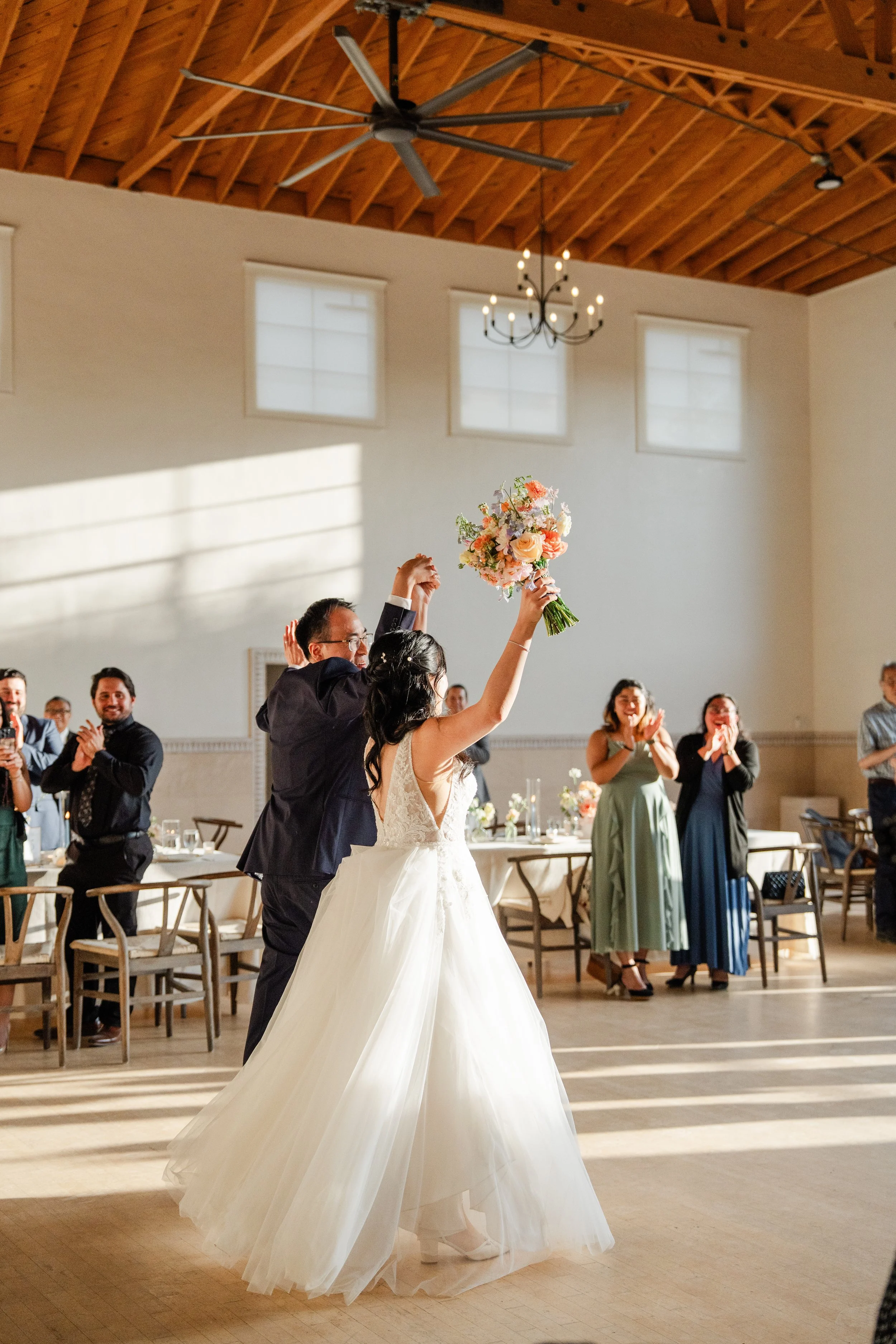 A bride in a white wedding gown holding a bouquet of flowers is dancing with a man in a tuxedo in a reception hall. Other guests are clapping and smiling in the background, with a high wooden ceiling, windows, and chandeliers overhead.