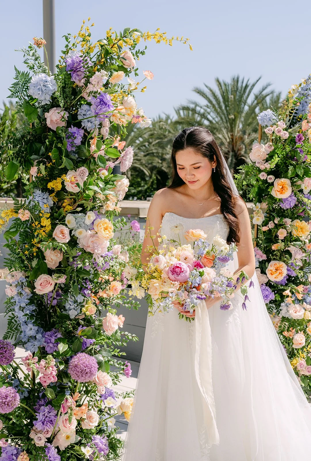 Linh looking down at her bouquet beside a lush floral arch