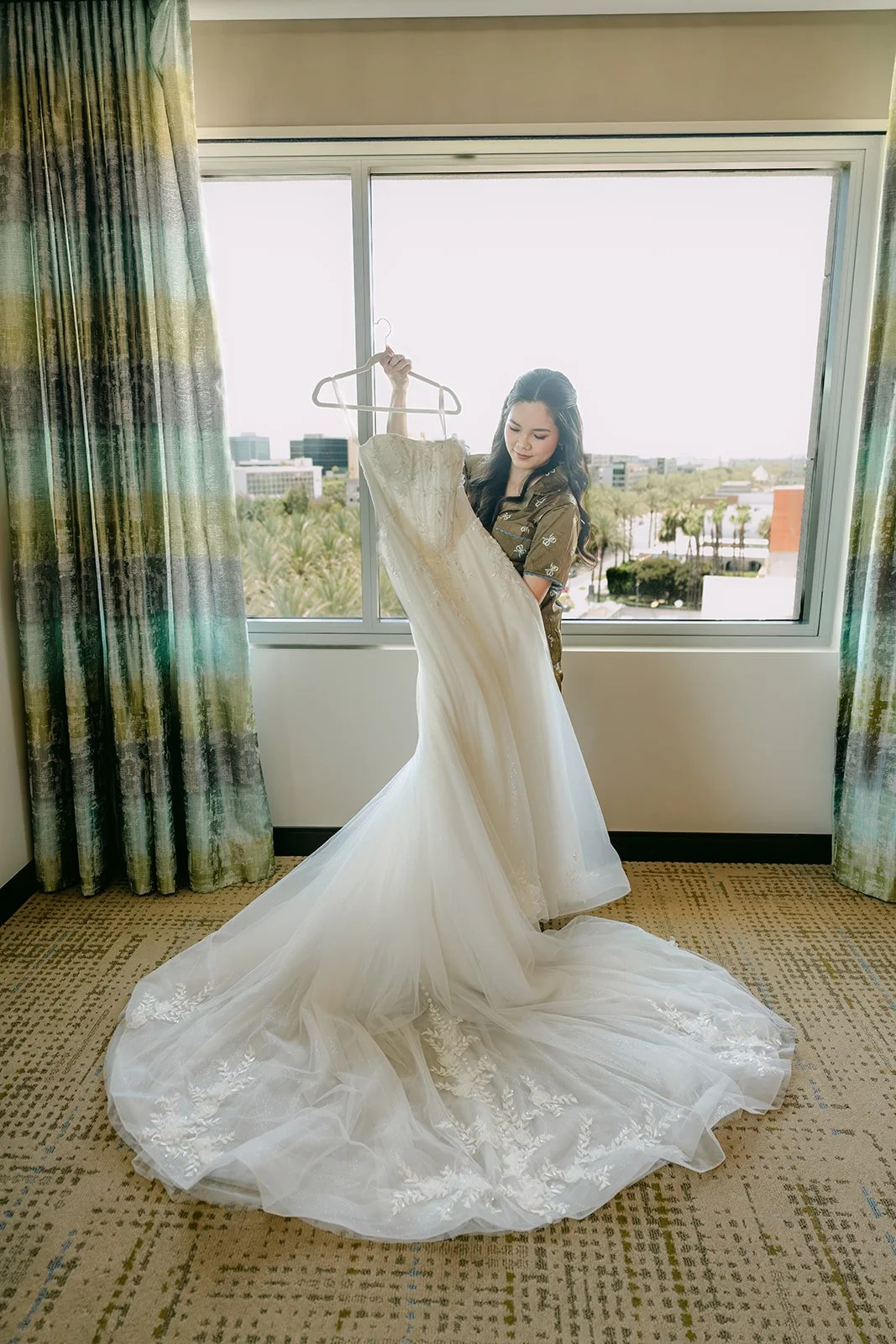Bride holding up her wedding gown by the window of her JW Marriott Anaheim suite