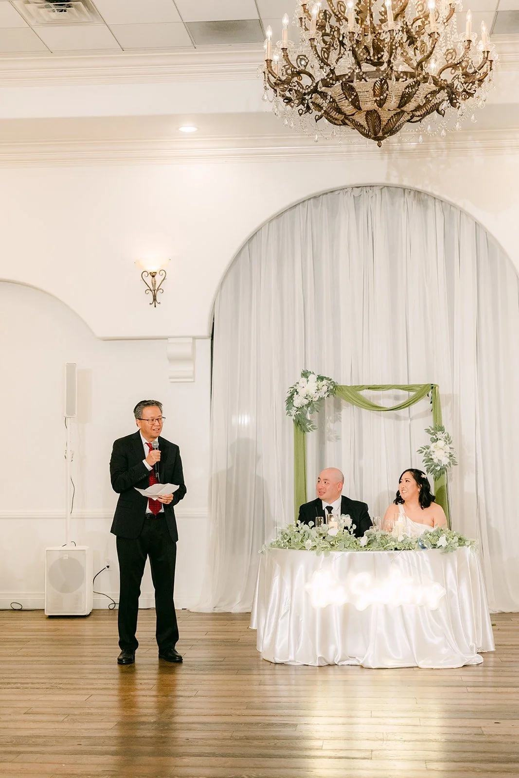 A speech beside the sweetheart table under the chandelier