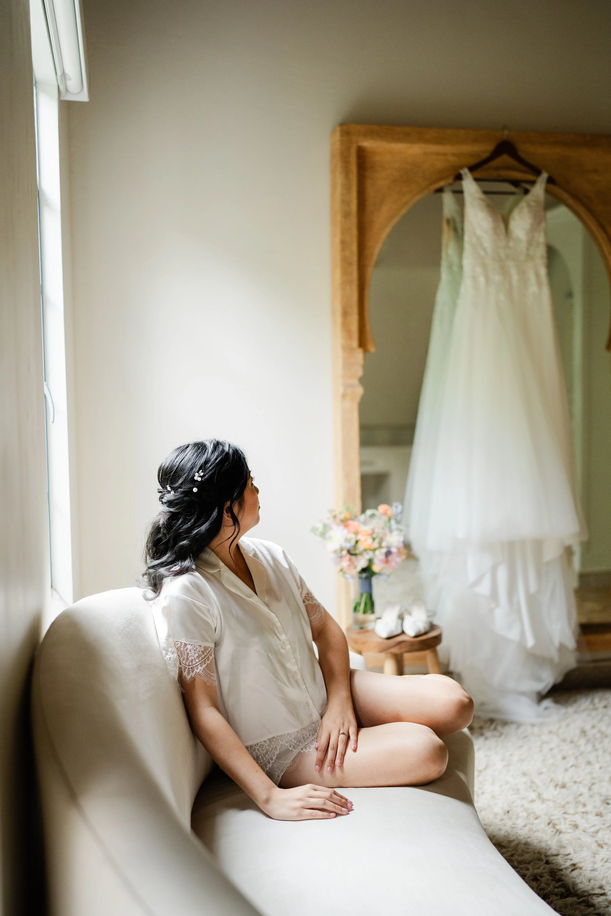 A woman with dark hair styled in loose waves, wearing a satin robe, sitting on a beige sofa in a softly lit room, looking towards a wedding dress hanging on a wooden frame in the background.