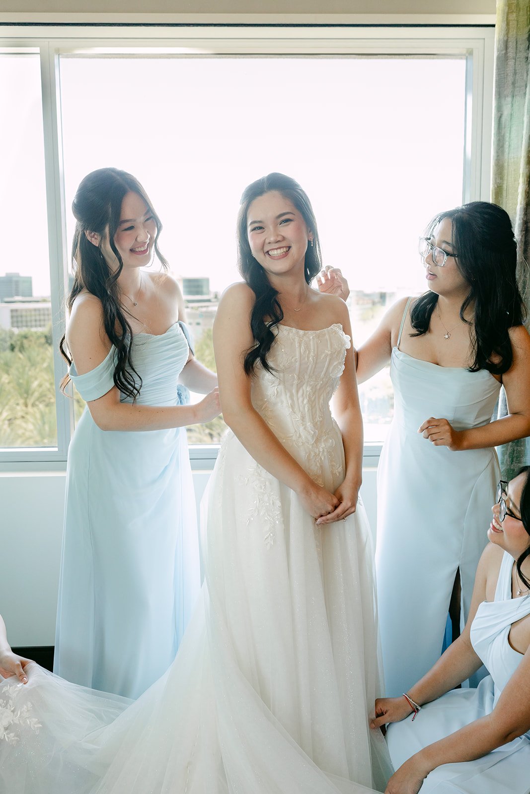 Bride laughing with her bridesmaids in light blue dresses beside the window