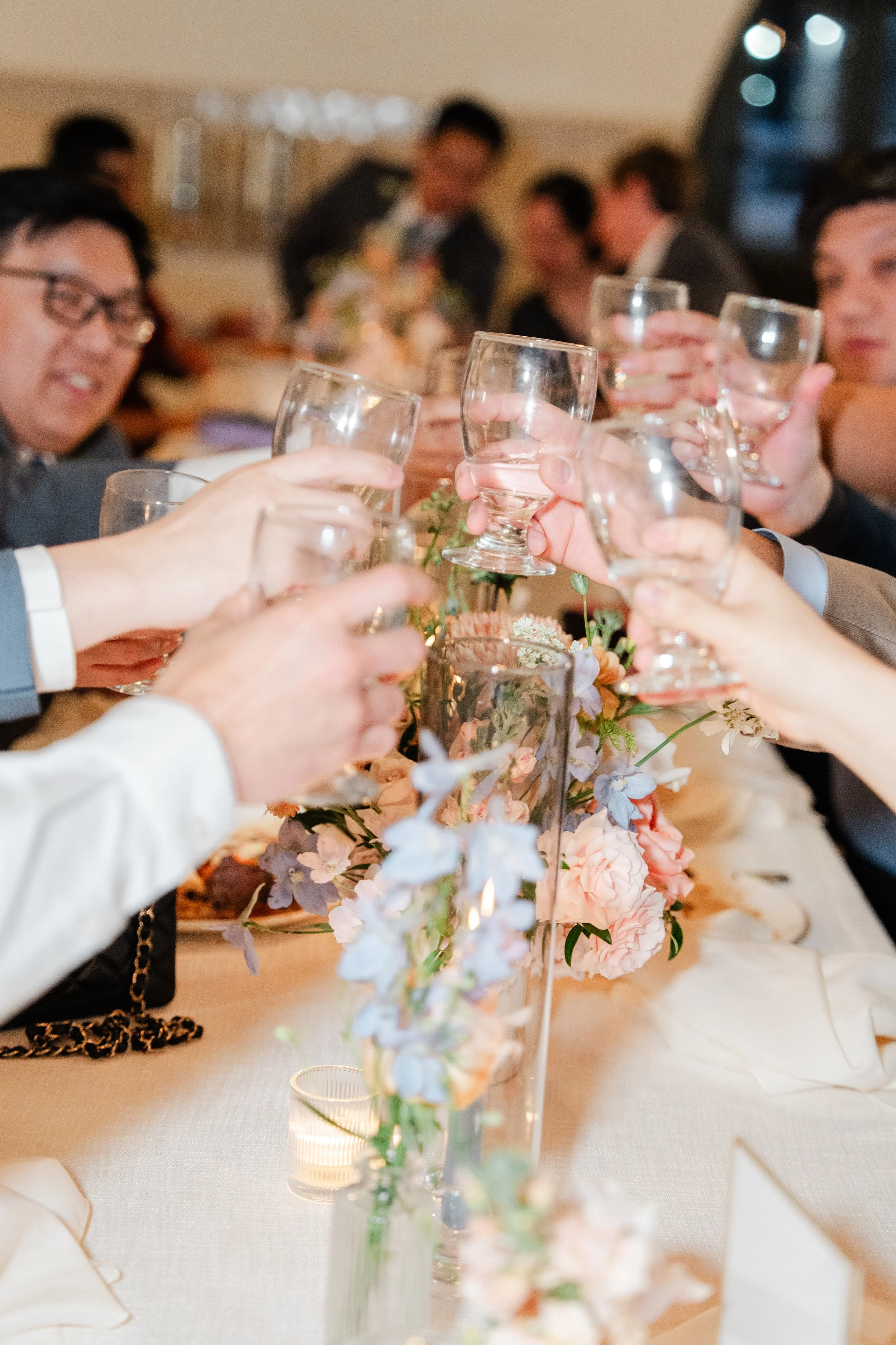 People raising glasses in a toast at a formal dinner with floral centerpiece.