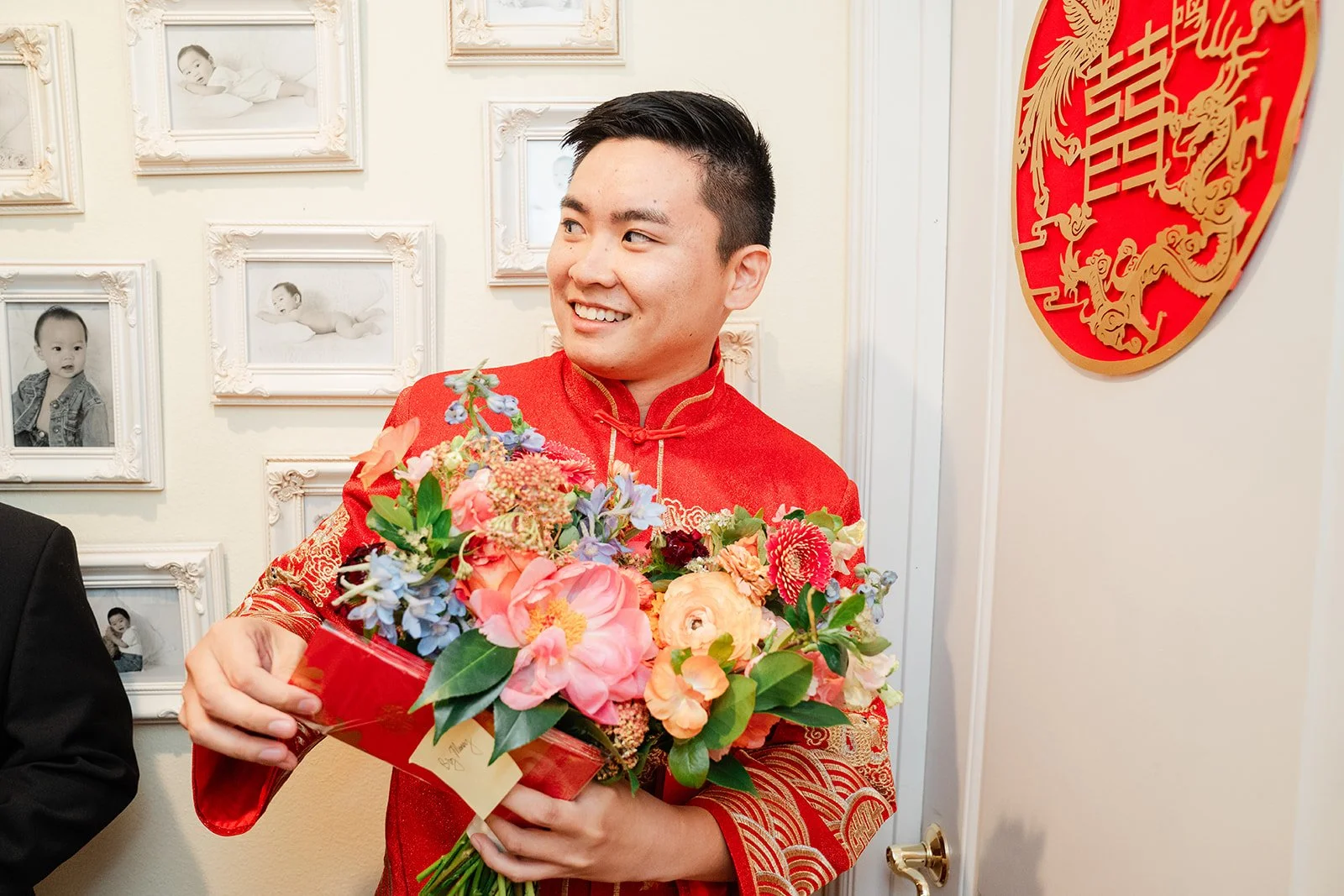 A groom in a red jacket holding the bride's bouquet before the door game.