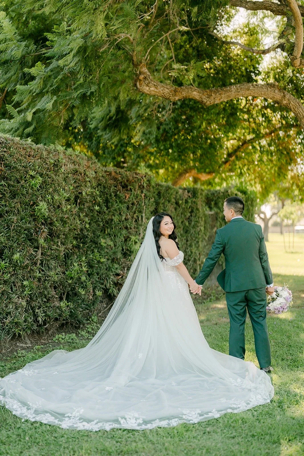 Jennifer and Kevin walk hand in hand along the hedge with her train behind