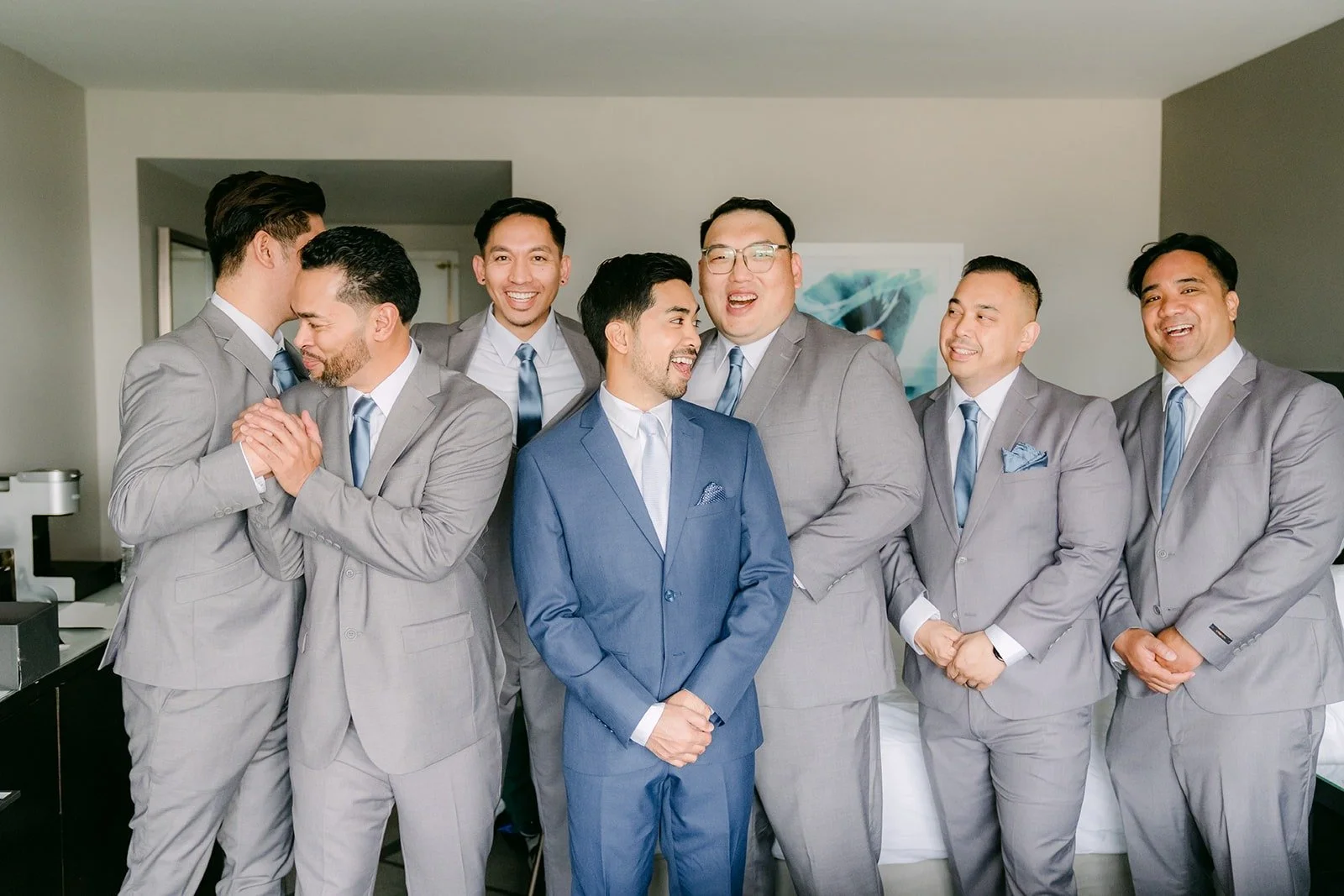 Mark in blue with his groomsmen in gray suits laughing together in the hotel room
