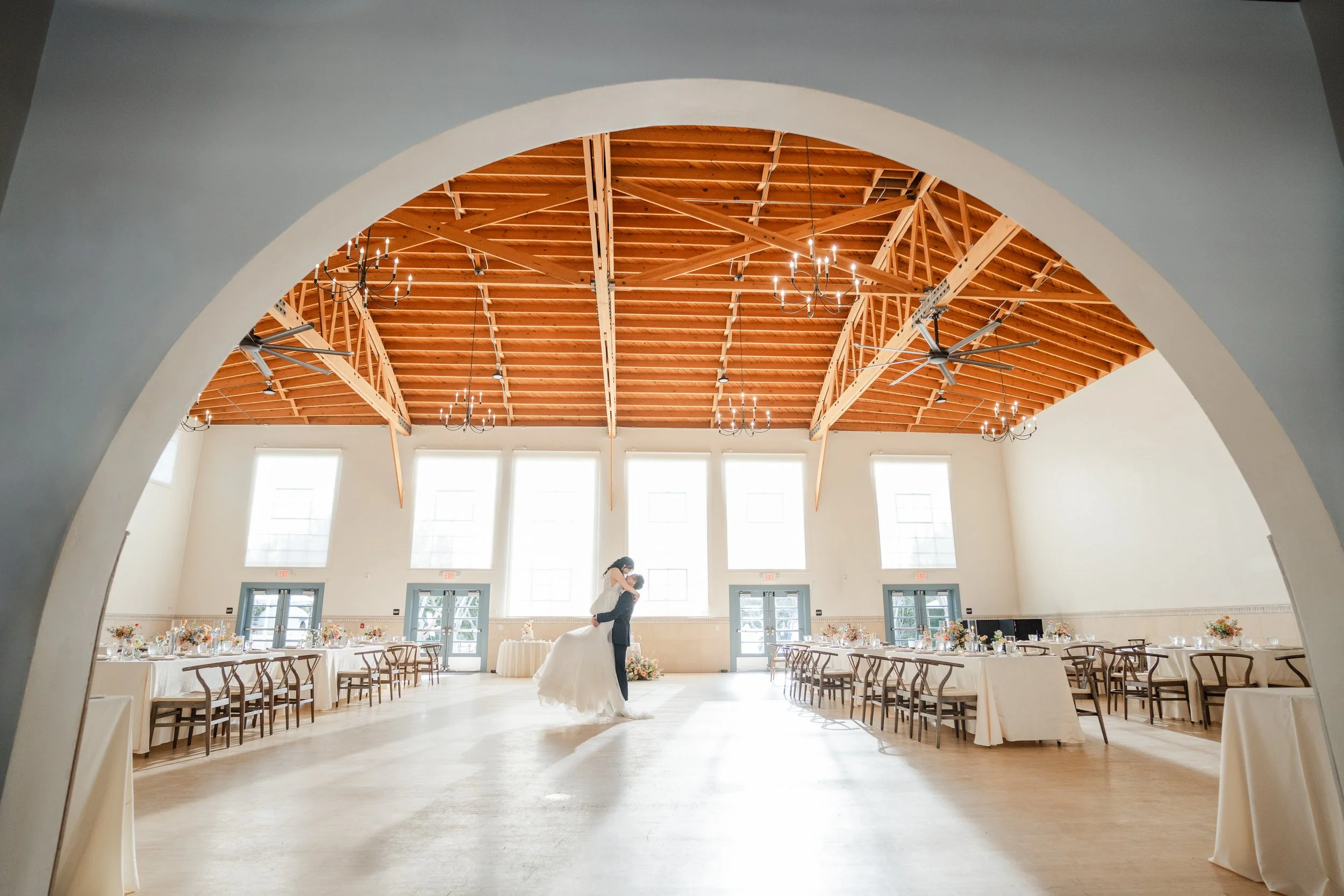 A bride and groom dancing in a large, bright wedding reception hall with high arched ceiling, wood beams, chandeliers, and tables decorated with flowers.