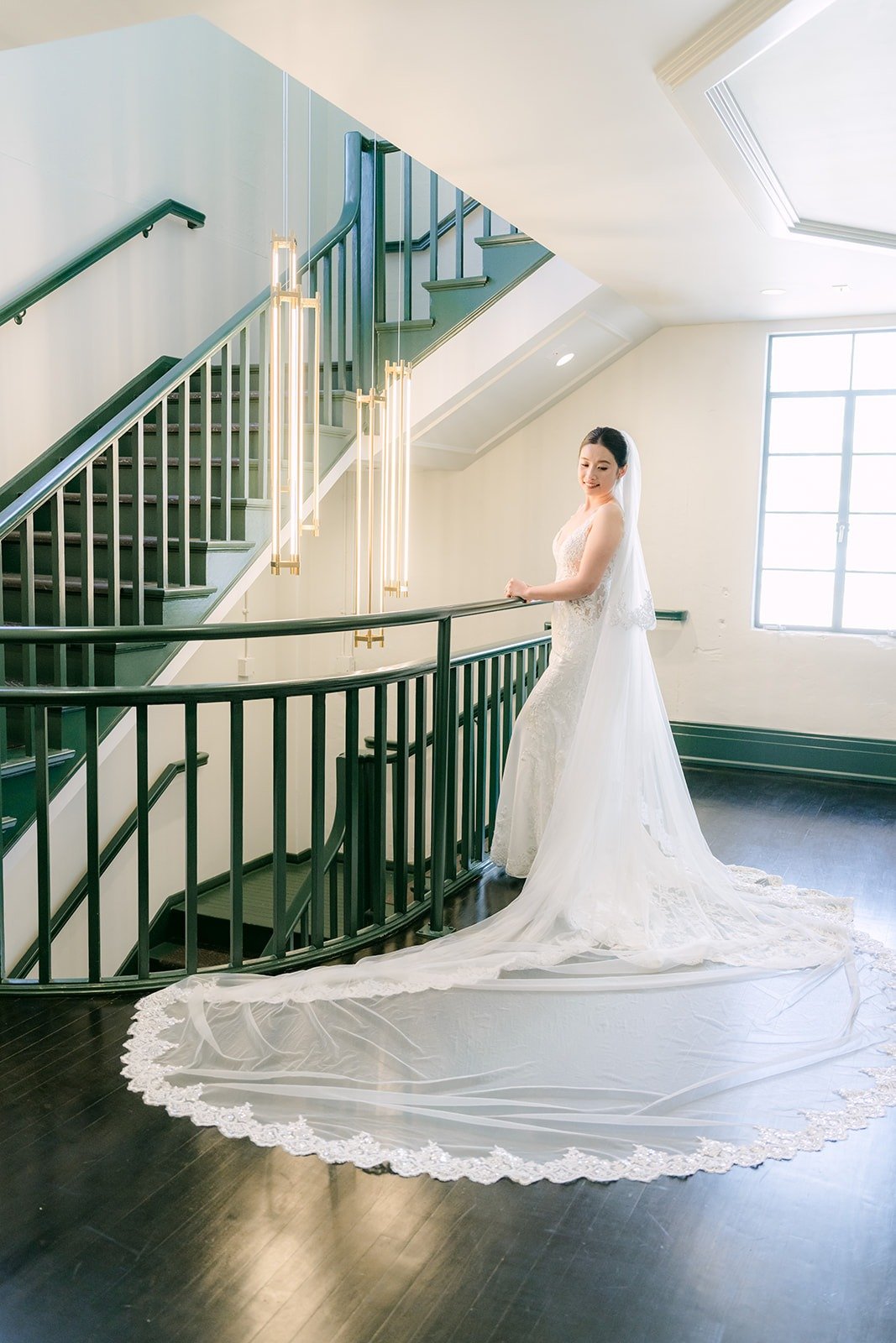 Elisa stands at the base of a green staircase with her lace train spread behind her
