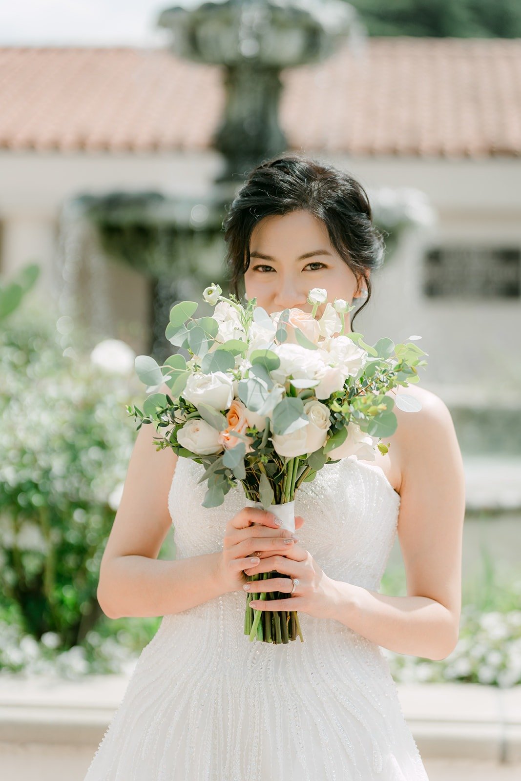 Eunice holds her bouquet of white and blush roses by the fountain at San Gabriel Mission