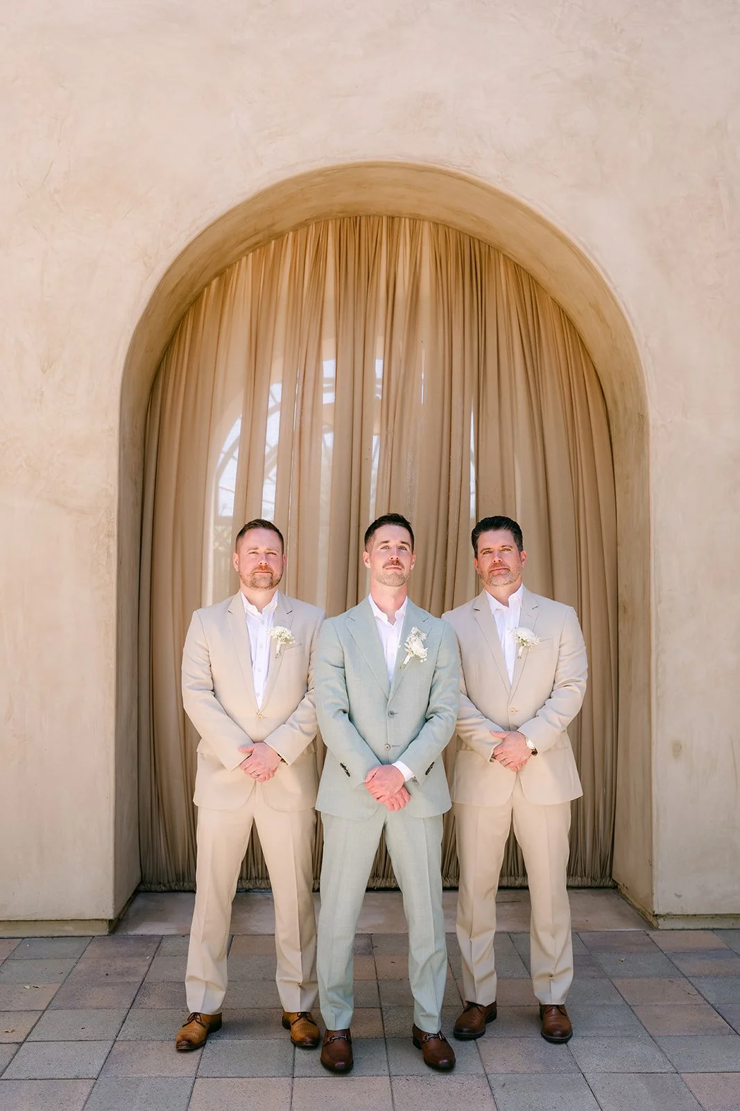 Anthony in a sage suit standing between two groomsmen under a curtained archway at Serra Plaza