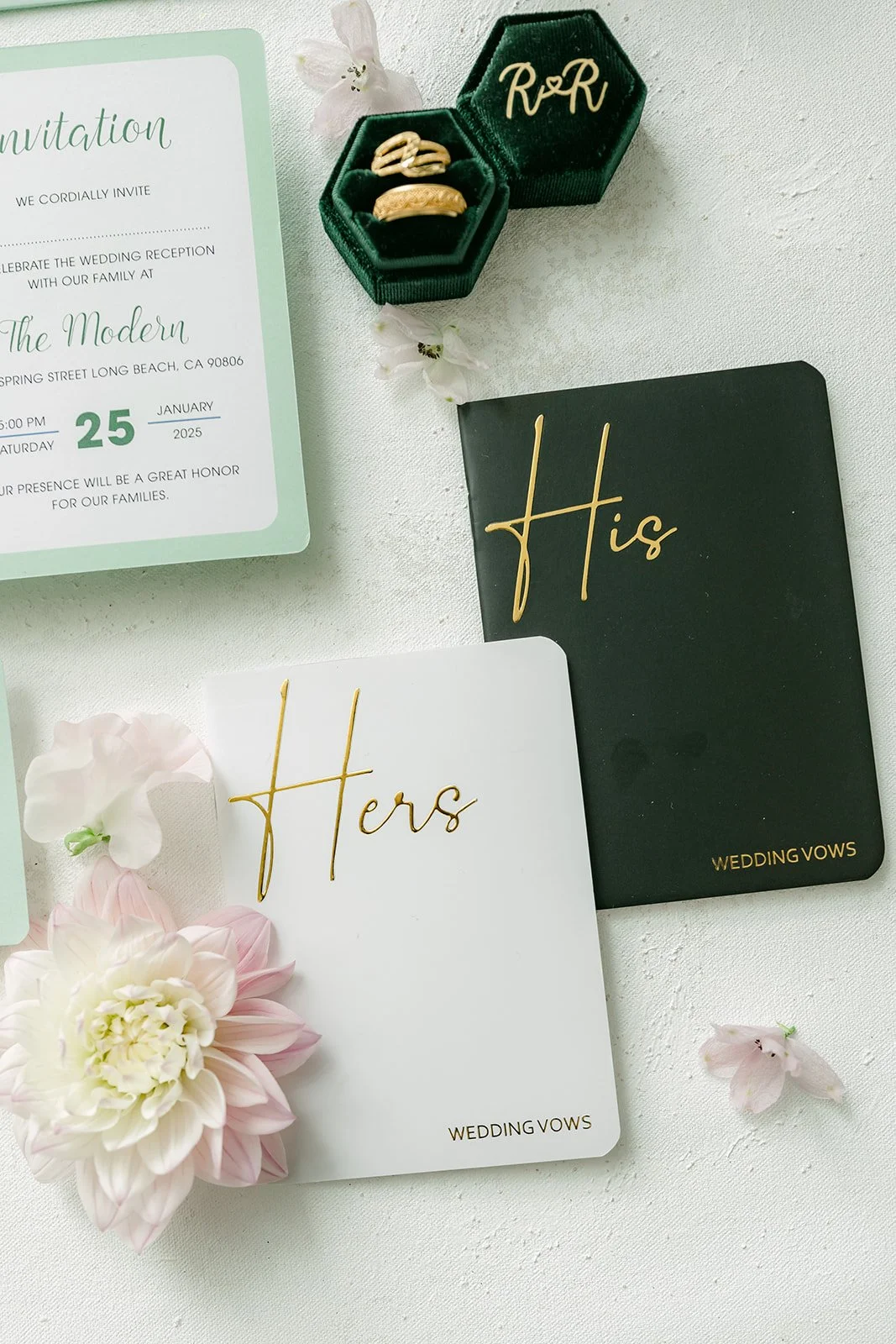 His and Hers vow books beside the invitation and green ring box with wedding bands