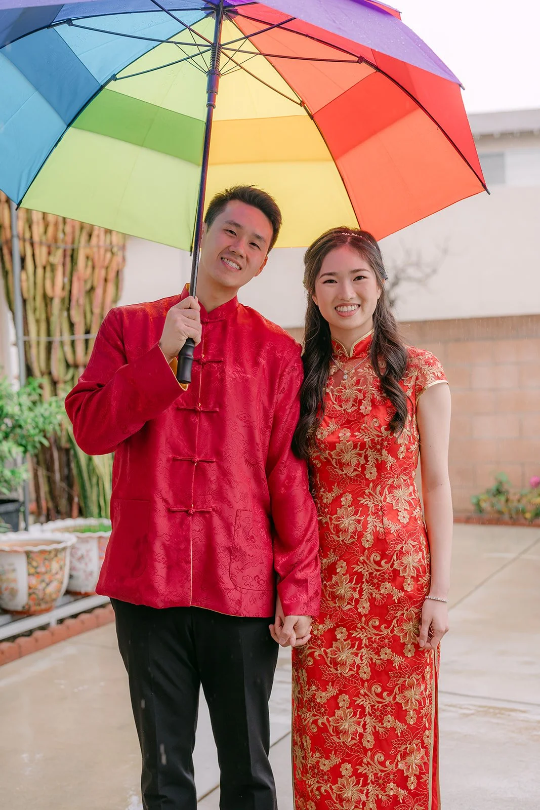 A couple in red standing under a rainbow umbrella in the backyard.