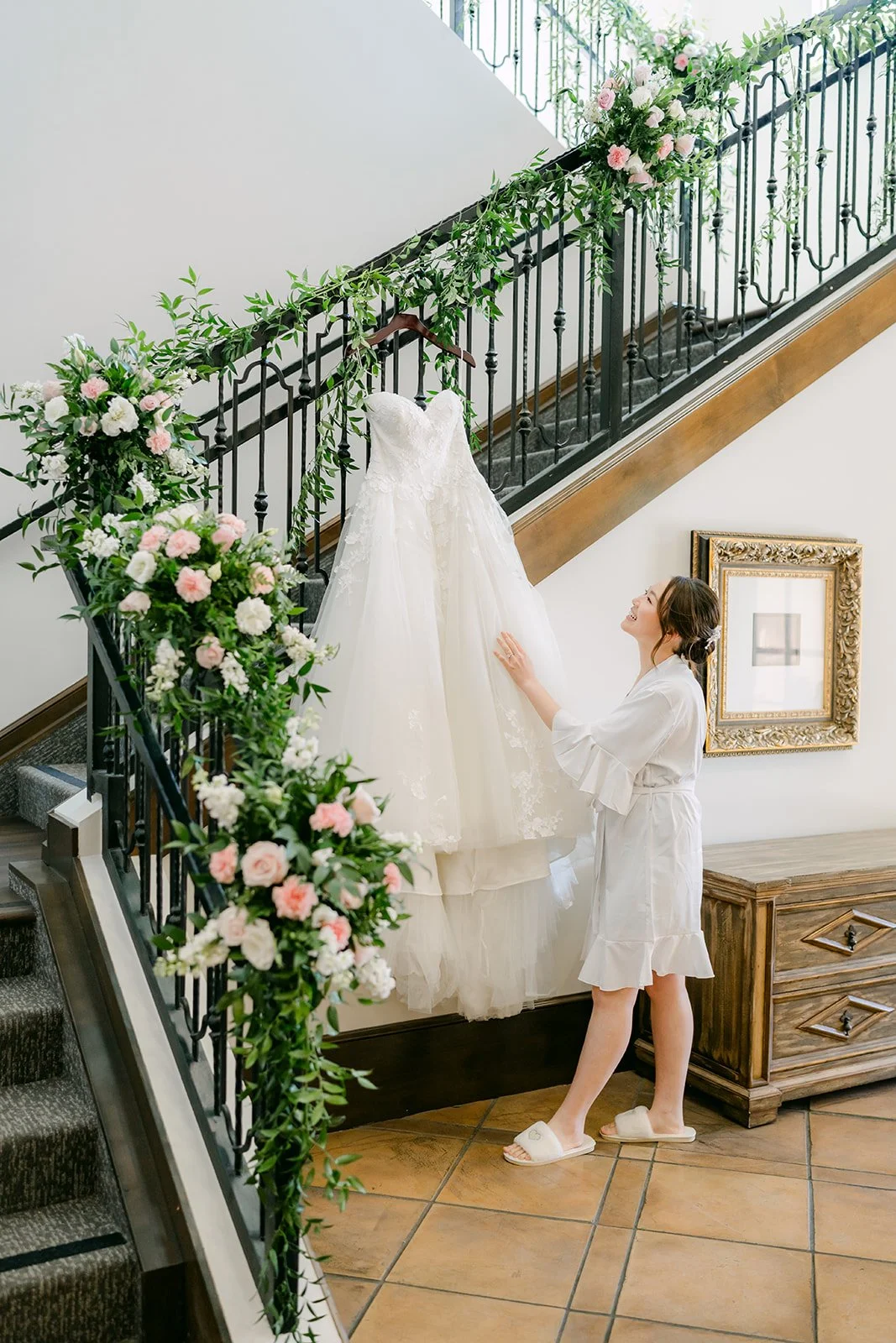 Bride in a white robe admiring her wedding gown hanging on the floral staircase at Vellano Estate