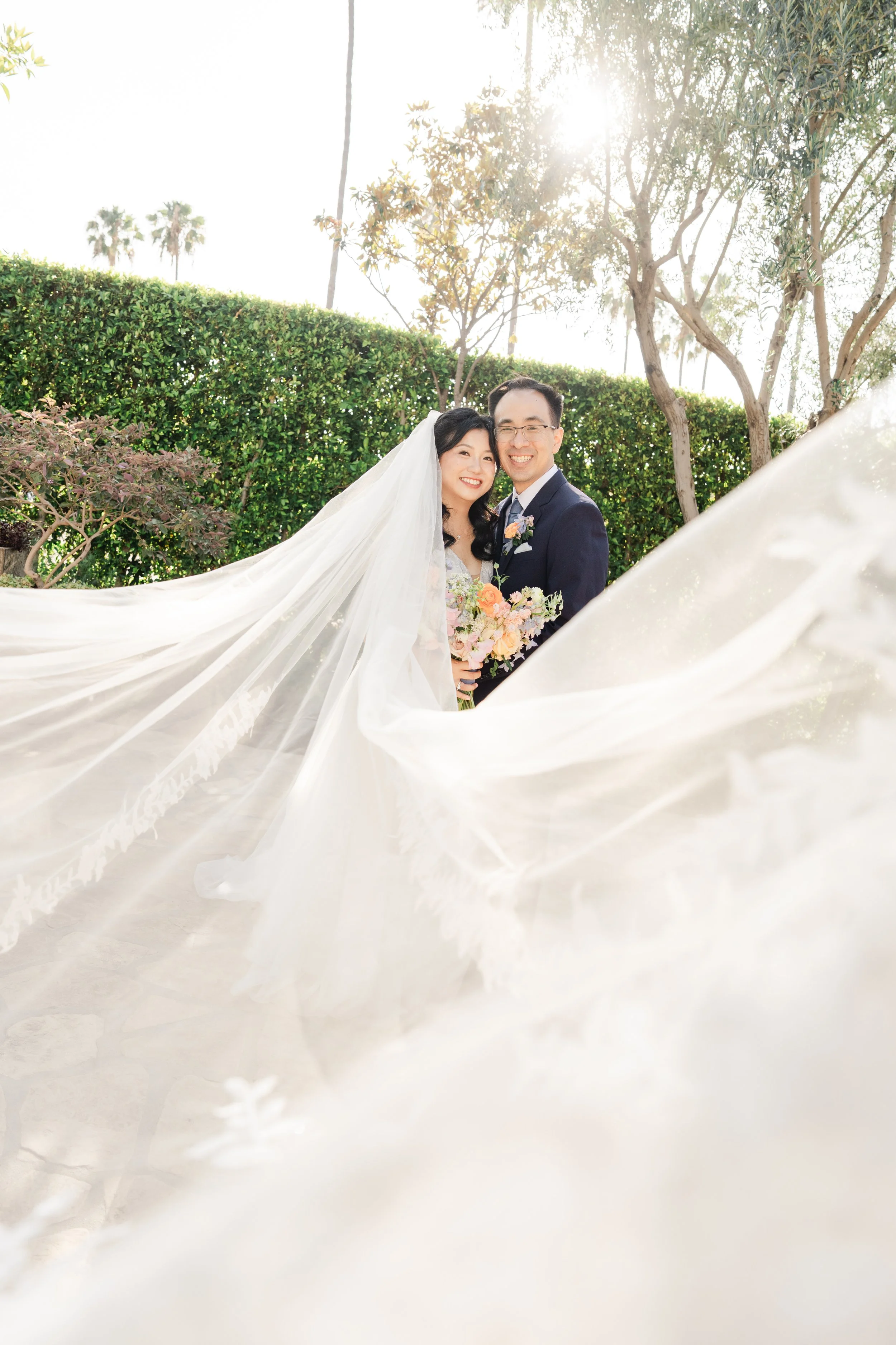 A newlywed couple smiling and embracing outdoors, with greenery and trees in the background. The bride is wearing a white wedding gown and veil, holding a bouquet, and the groom is wearing a navy suit. The sunlight filters through the trees, creating