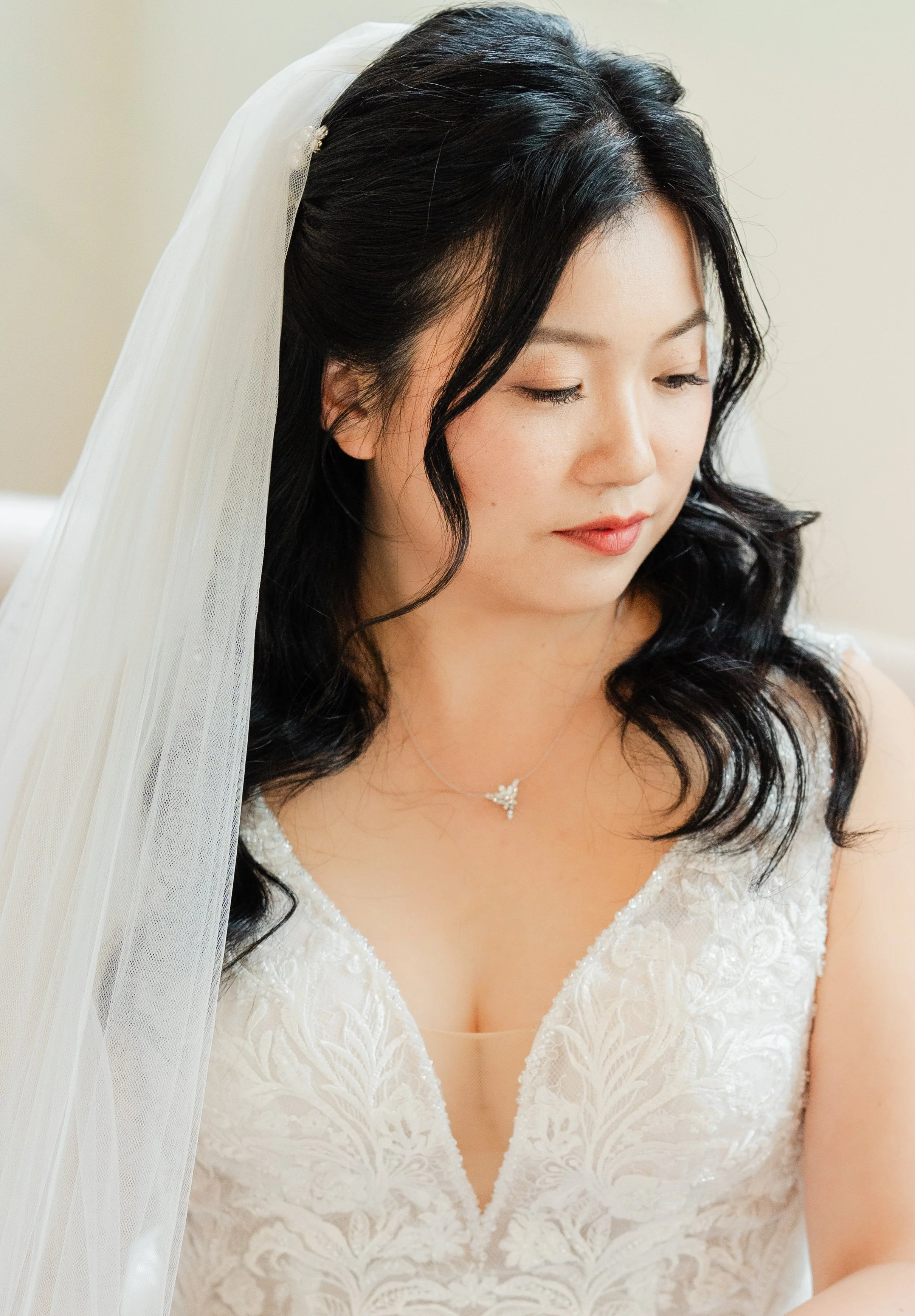 Close-up of a woman with black wavy hair wearing a white wedding dress with lace detail and a veil, looking downward.