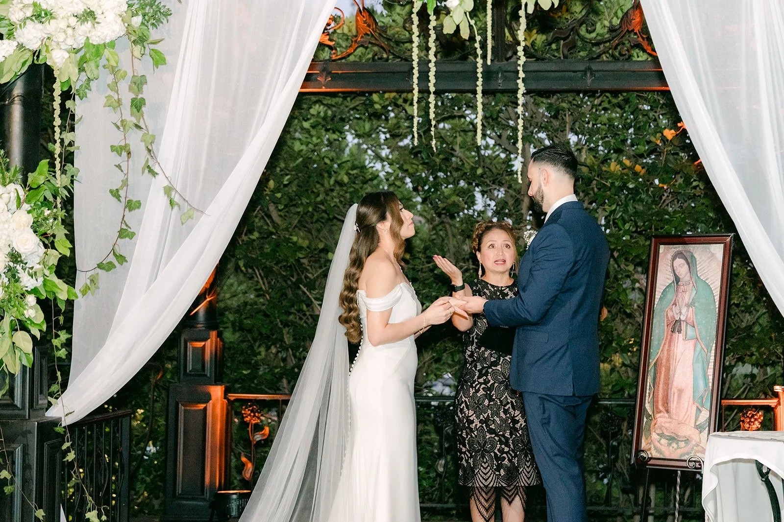 Mariana and Danny exchanging rings with their bilingual officiant beside an image of the Virgin Mary