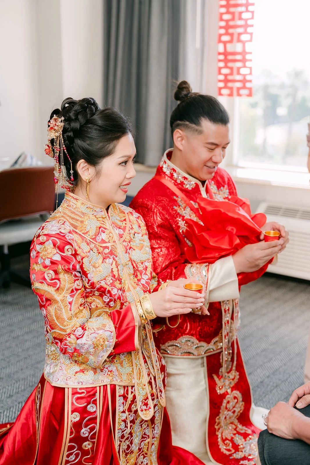A bride and groom in red holding teacups as they wait to serve the next relative.