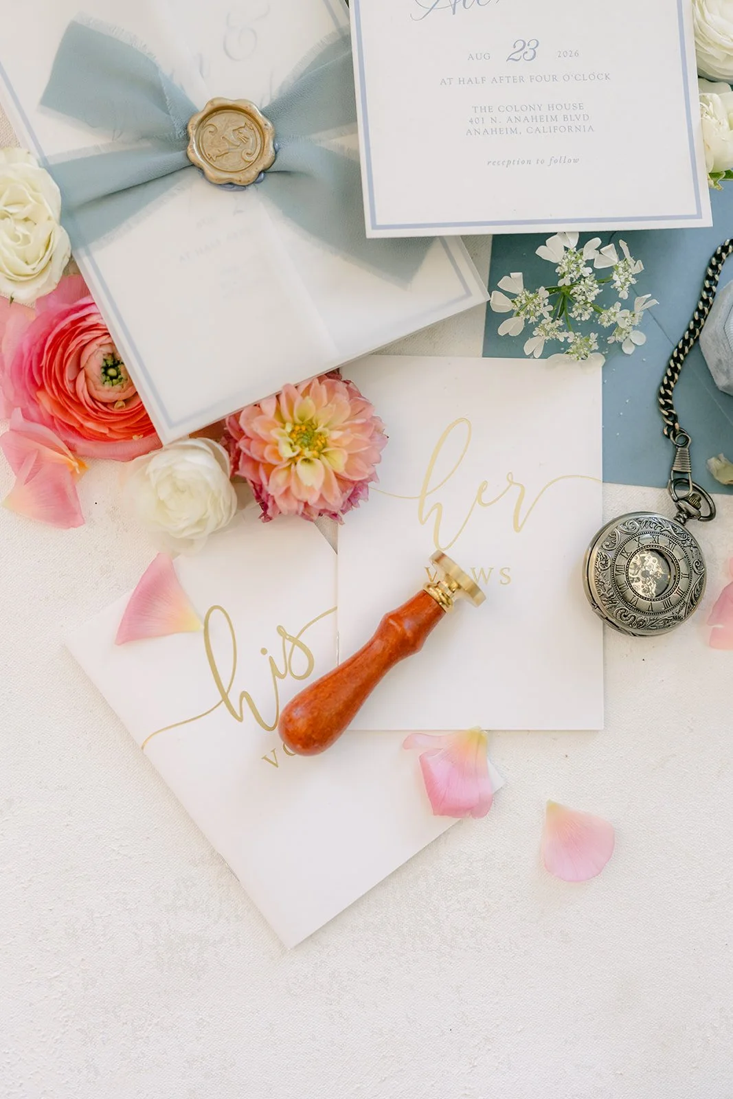 Flat lay with a dusty blue invitation suite, wax seal, ribbon and pink ranunculus